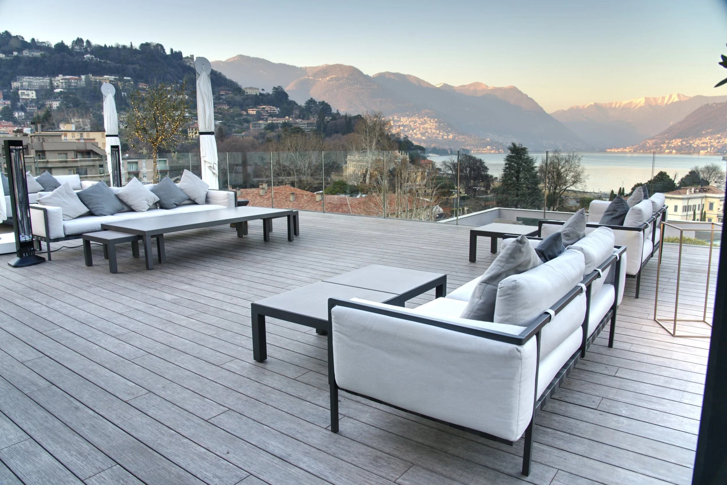 A spacious outdoor patio with modern furniture and panoramic views of mountains, water, and a hilly cityscape.