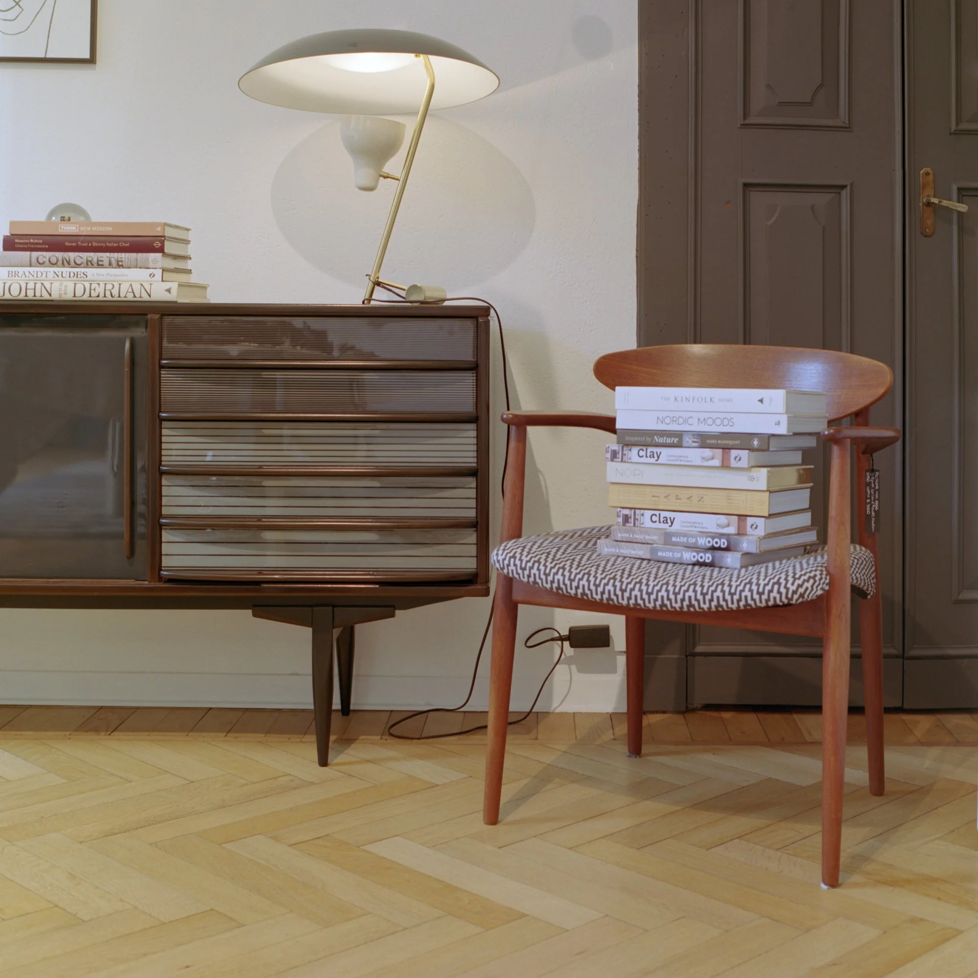 A wooden chair with a stack of books on it, a wooden cabinet, a white wall, a dark wooden door, and a modern lamp on top of the cabinet.