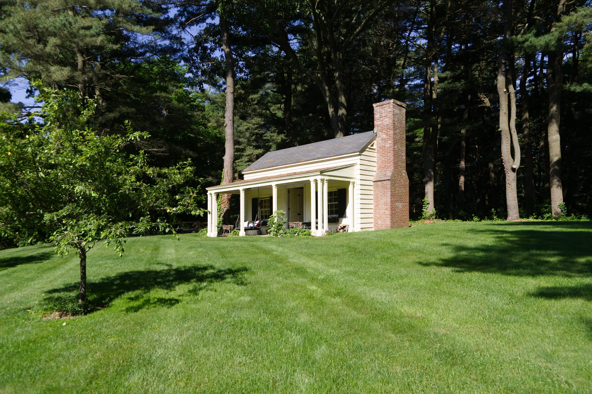 A house with a covered porch, white siding, brick chimney, and gazebo in a lush green yard surrounded by tall trees.