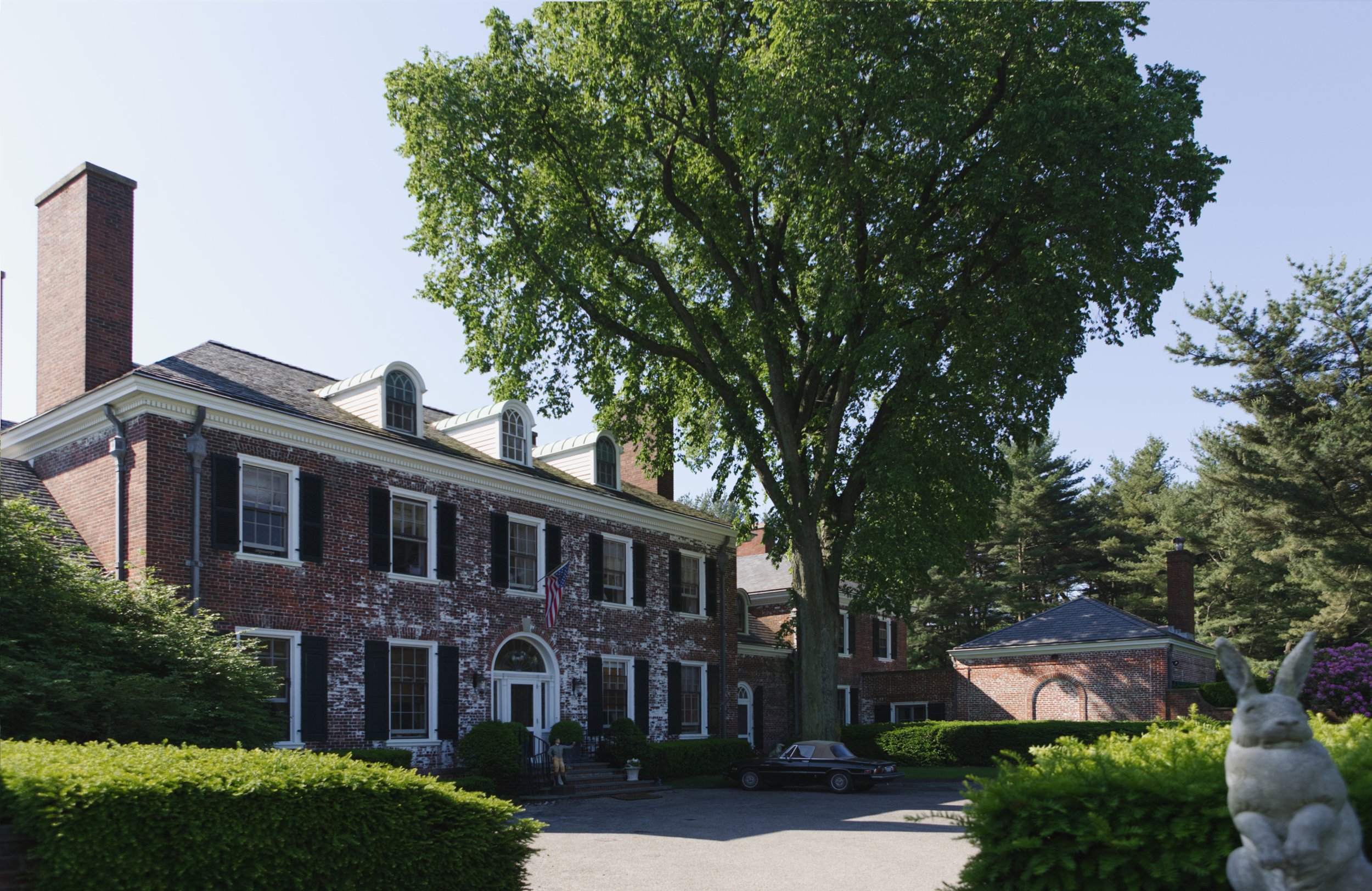 A large brick house with white trim, multiple windows with black shutters, a prominent chimney, and a front porch with steps. The house is surrounded by lush green trees and bushes. A black car is parked in the driveway. The scene is bright and sunny