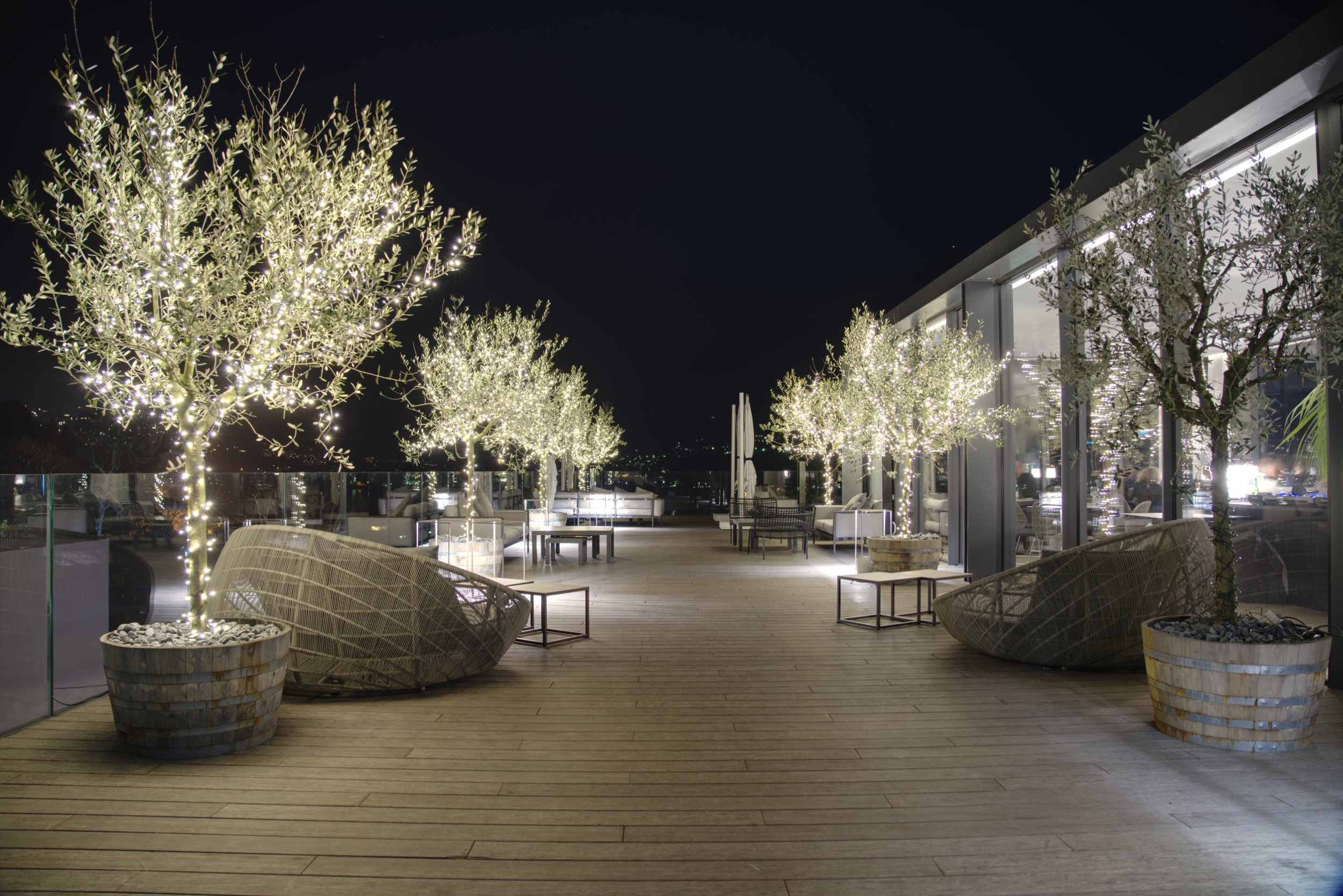 Outdoor patio at night decorated with illuminated string lights on trees, with modern furniture, potted plants, and a glass railing overlooking a cityscape.