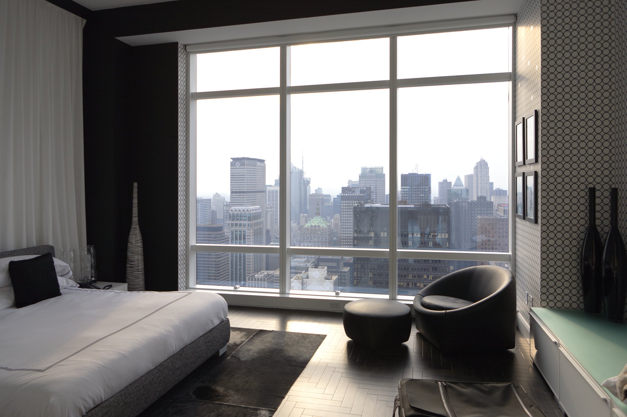 Modern bedroom with large floor-to-ceiling windows offering a city skyline view, a bed with black and white pillows, a black lounge chair, and geometric wallpaper on the wall.
