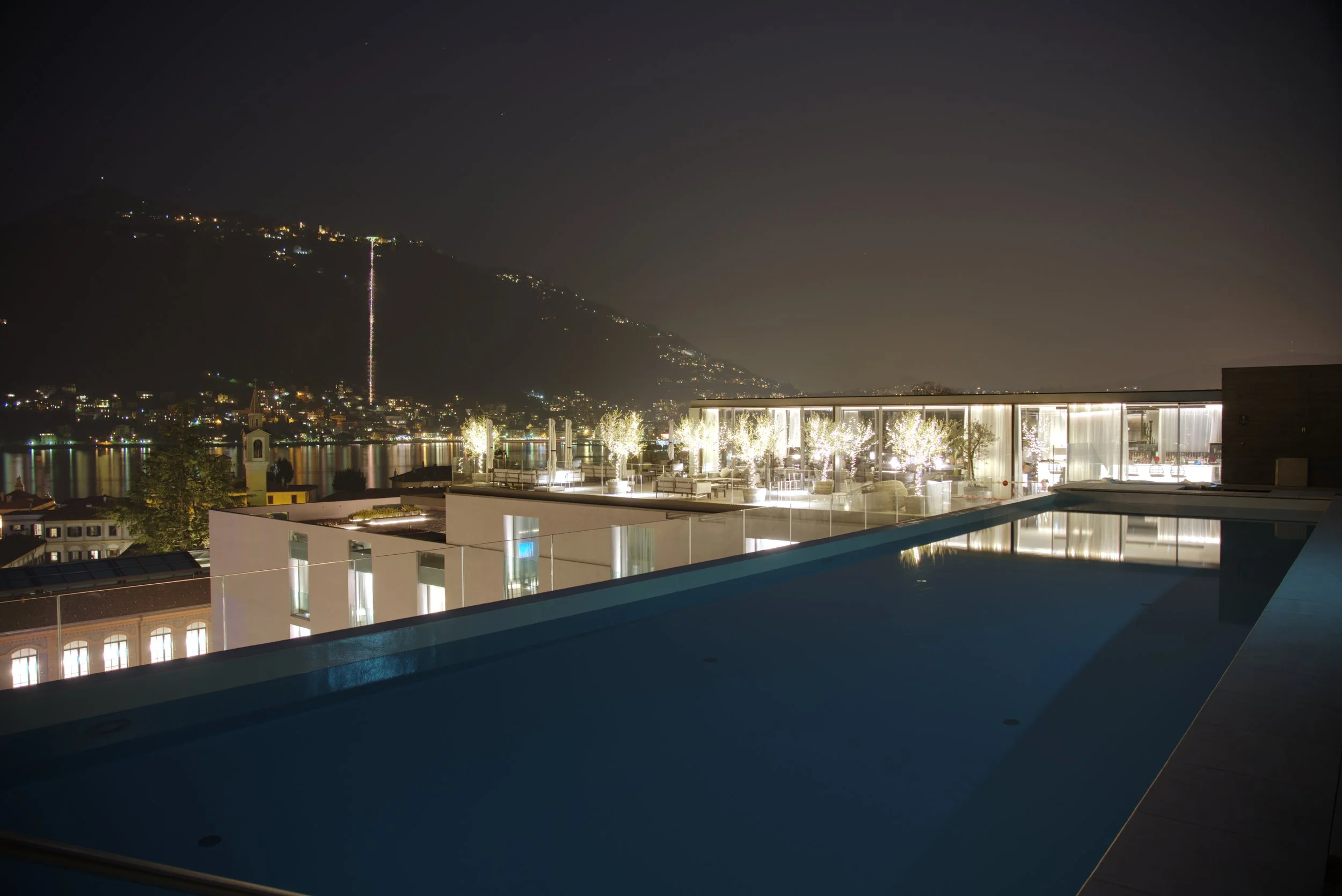 Night view of a city with a hill in the background, a lit-up tower, and a modern building with outdoor seating and decorative lights in the foreground.