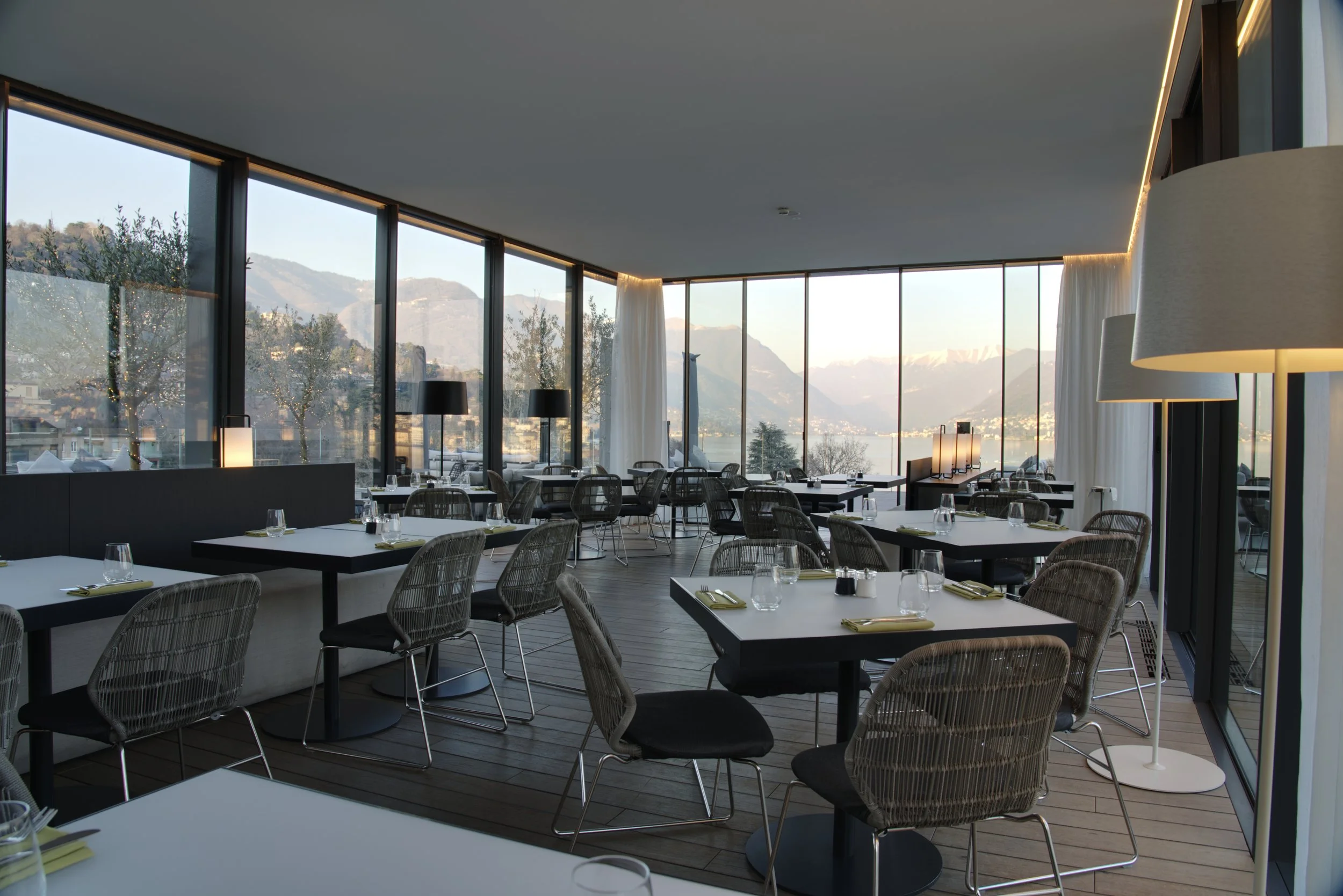 Modern restaurant with large floor-to-ceiling windows showing mountain views, arranged with tables, chairs, and minimal decor, illuminated by natural light.