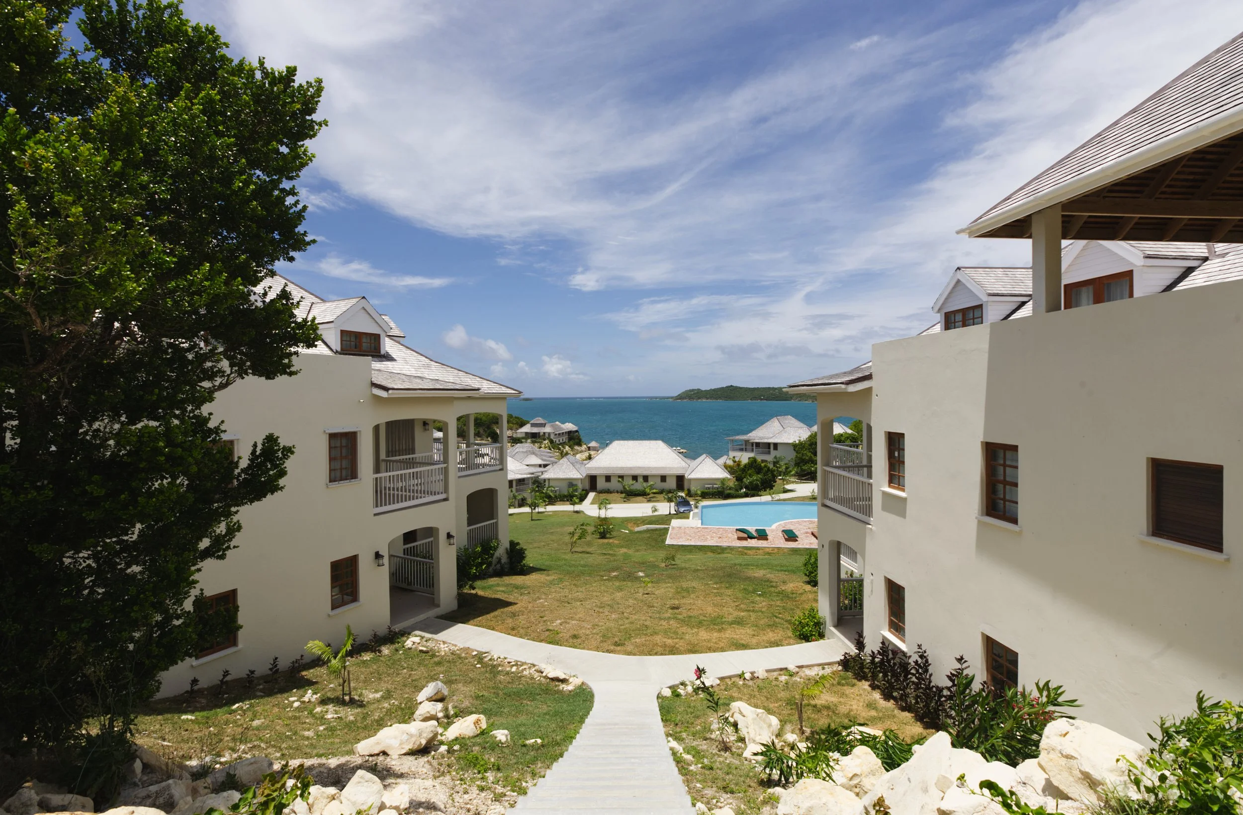 View of a luxury seaside resort with white buildings, green lawn, swimming pool, and ocean in the background under a partly cloudy sky.