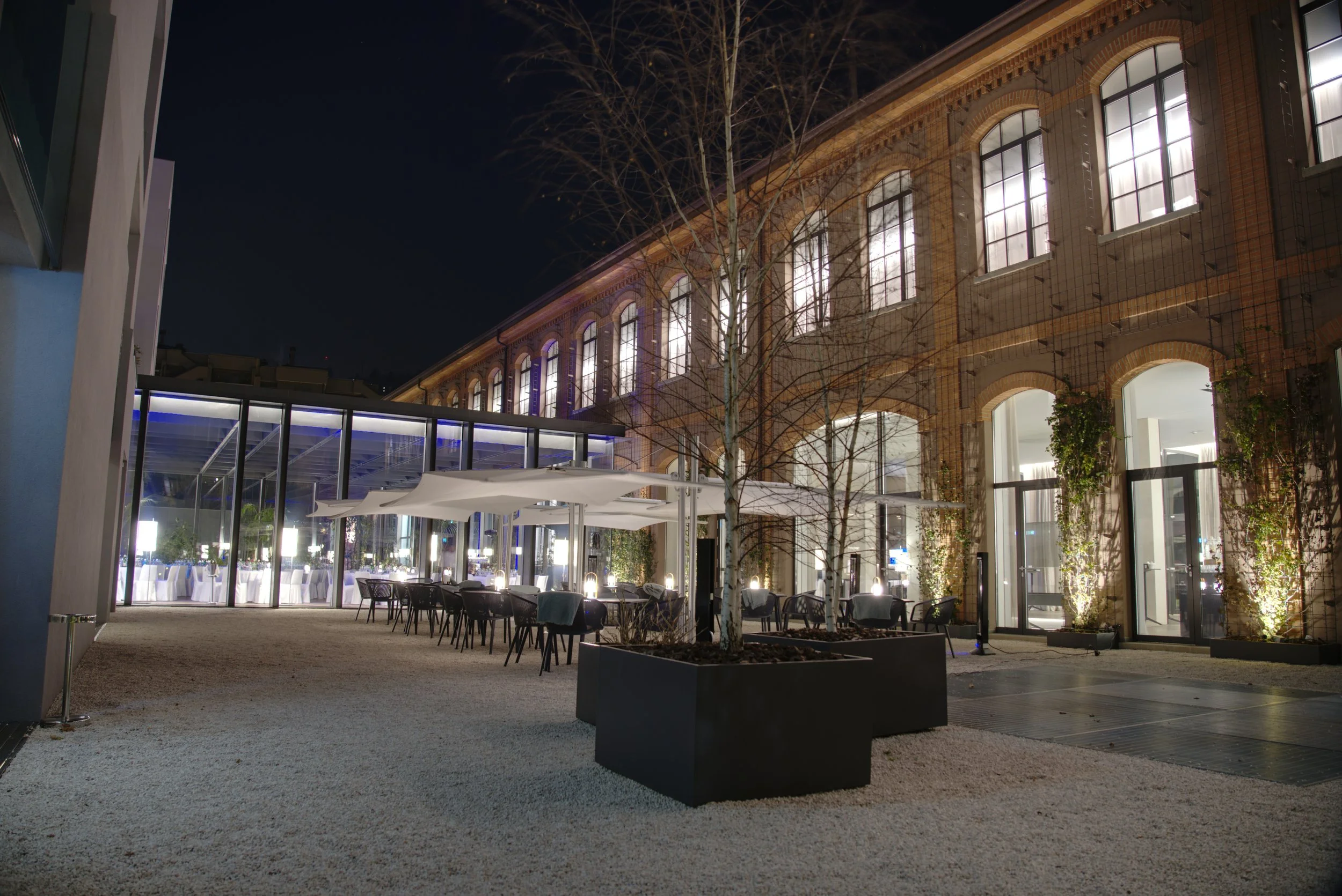 Night view of an outdoor dining area with tables and chairs, large potted plants, leafless trees, and a multi-story brick building with illuminated windows, seen from inside a building with glass walls and a glass ceiling.