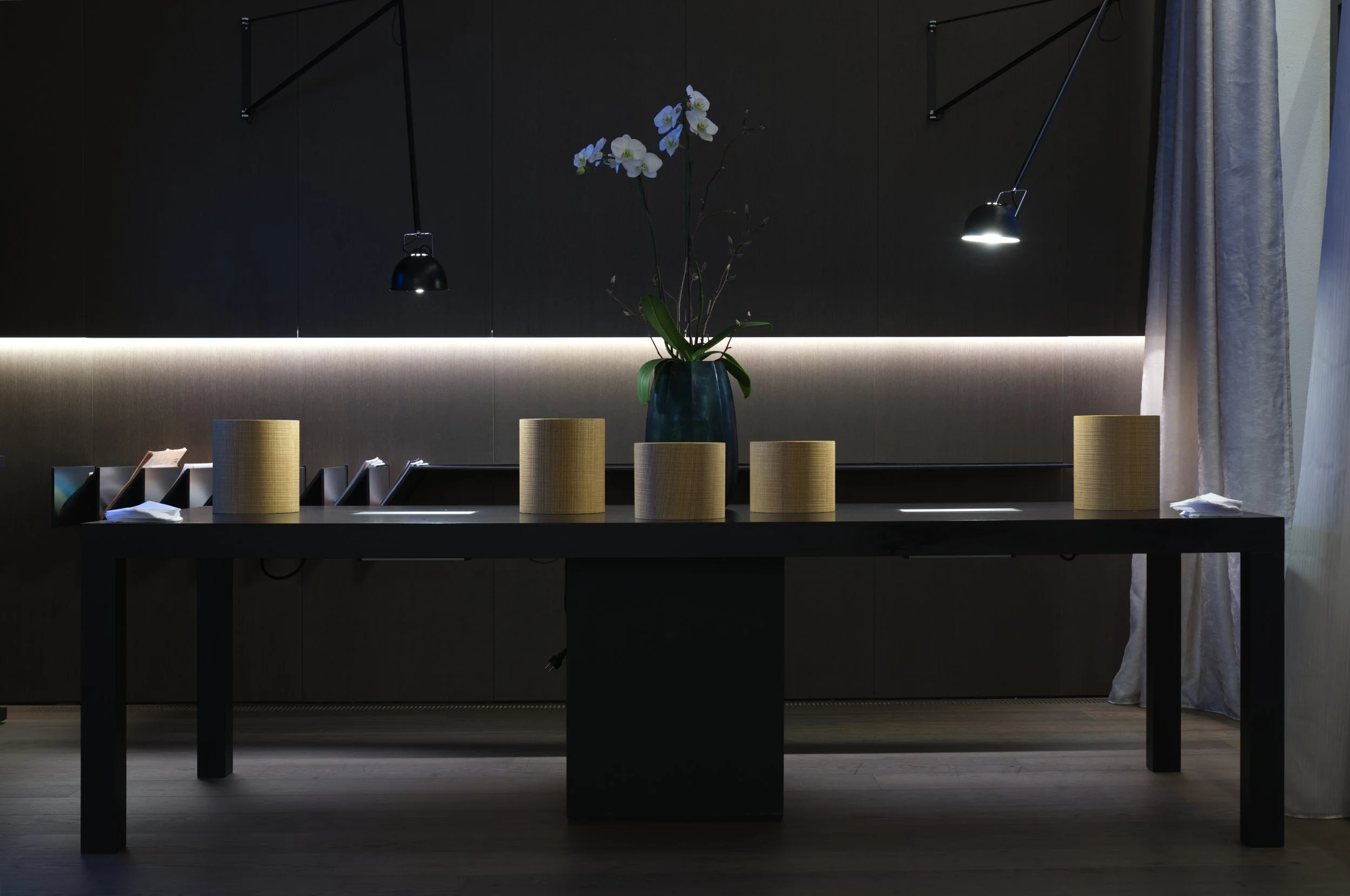 Modern indoor space with a curtain on the right, a dark wall, and a long table with a black frame and a dark top. On the table, there are stacks of white papers and three beige cylindrical objects. A blue vase with white orchids with green leaves is 