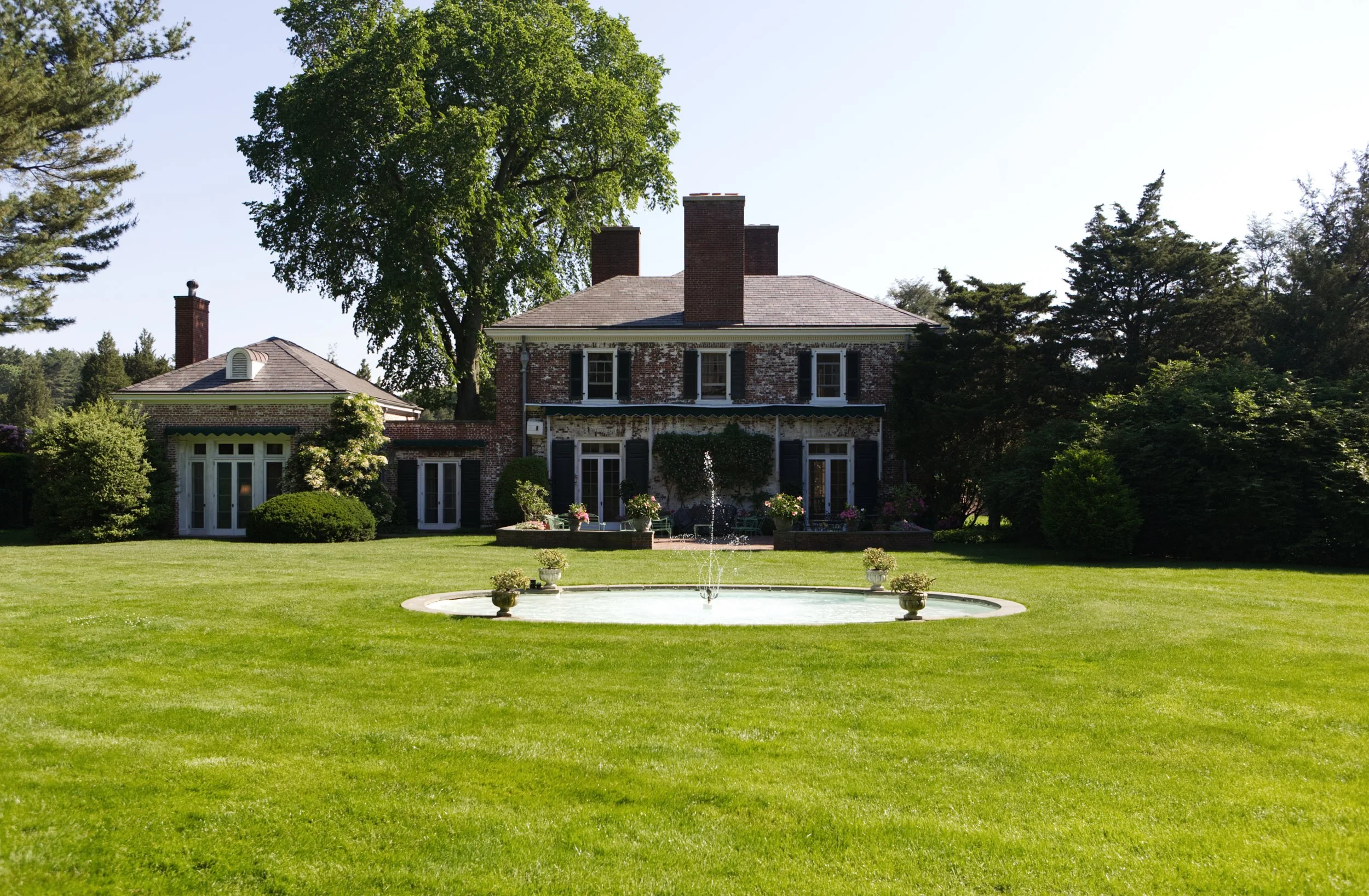 A large brick house with multiple chimneys, surrounded by lush greenery, trees, and a well-maintained lawn with a circular fountain in the foreground.