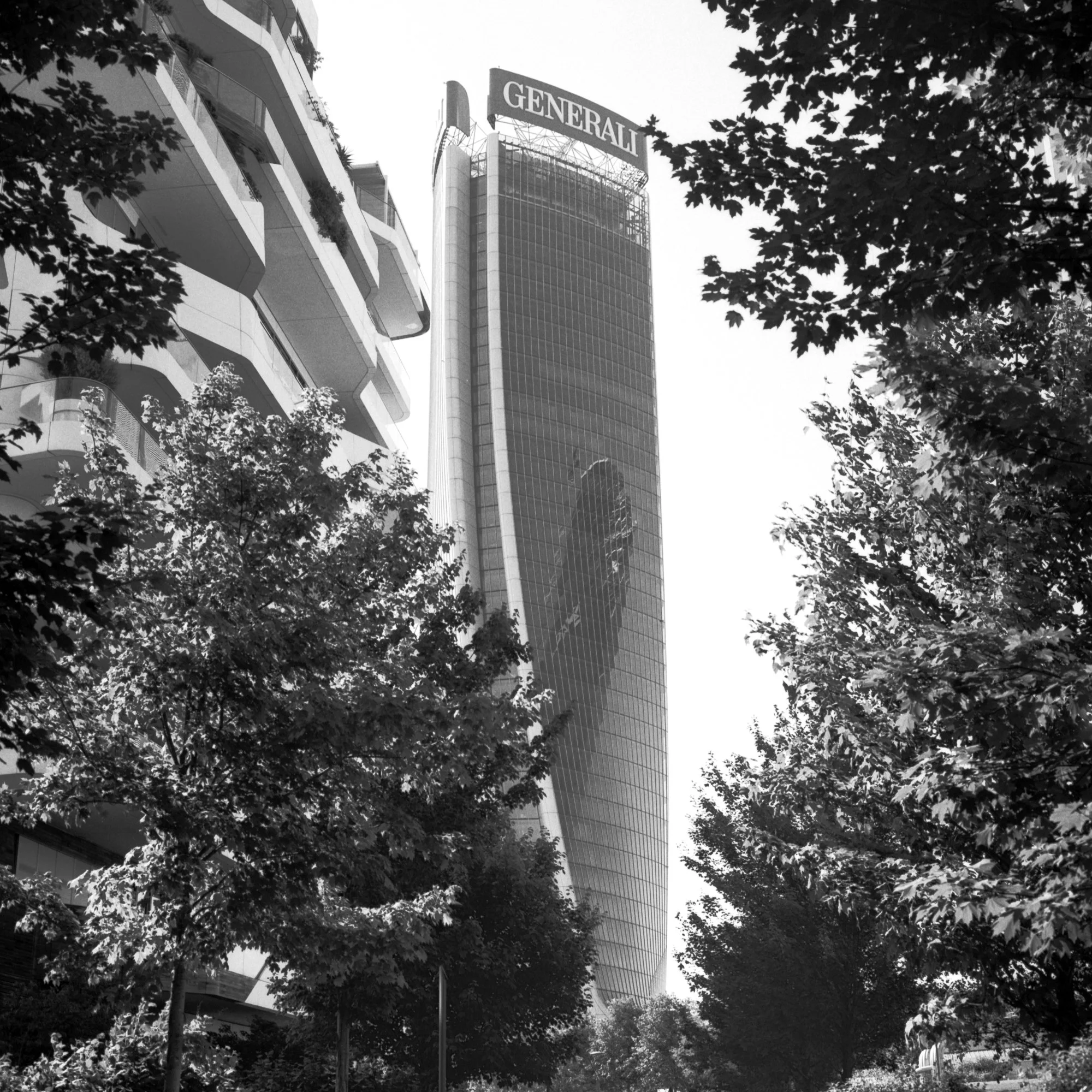 Black and white photo of a tall modern skyscraper with a large "GENERALI" sign on top, surrounded by trees in the foreground.