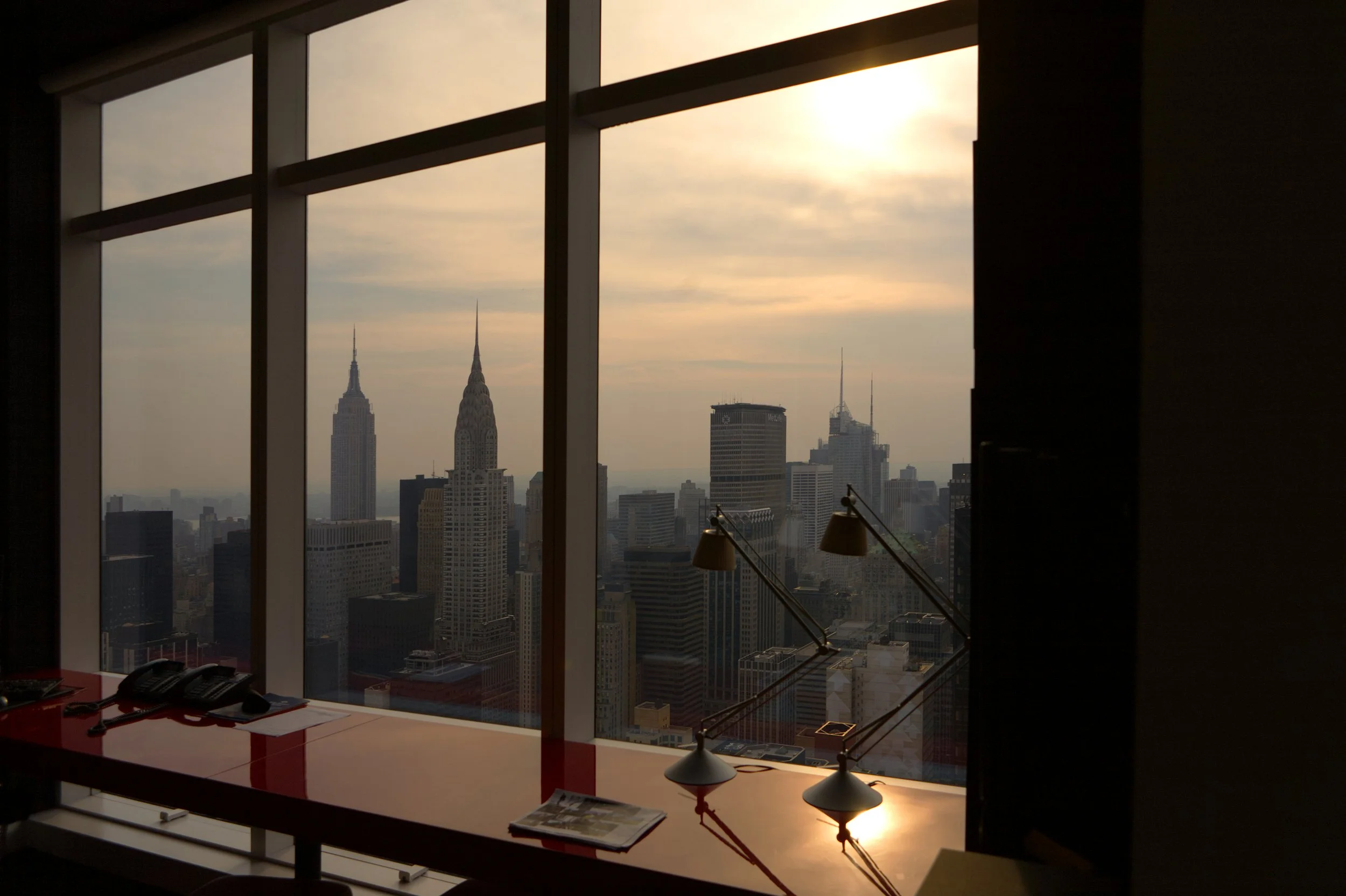 City skyline with tall buildings, including the Empire State Building and Chrysler Building, seen through office window during sunset.