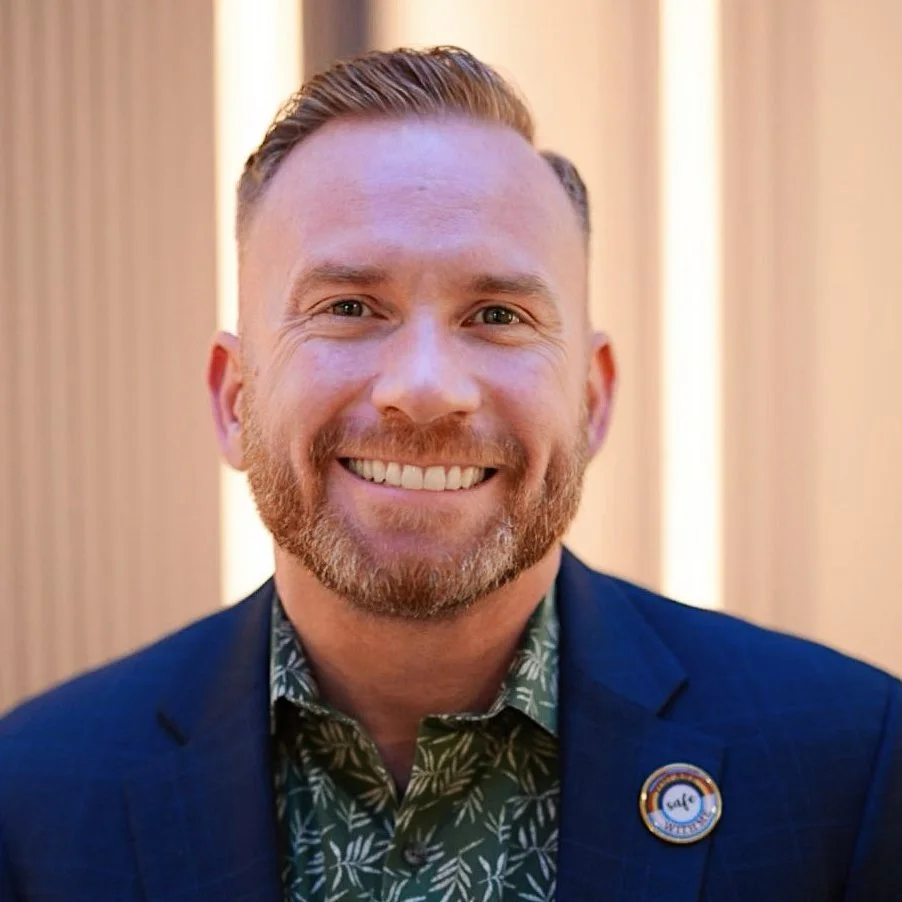 A smiling man with light skin, short reddish hair, and a beard, wearing a dark blazer and a patterned shirt, standing in front of a cream-colored curtain.