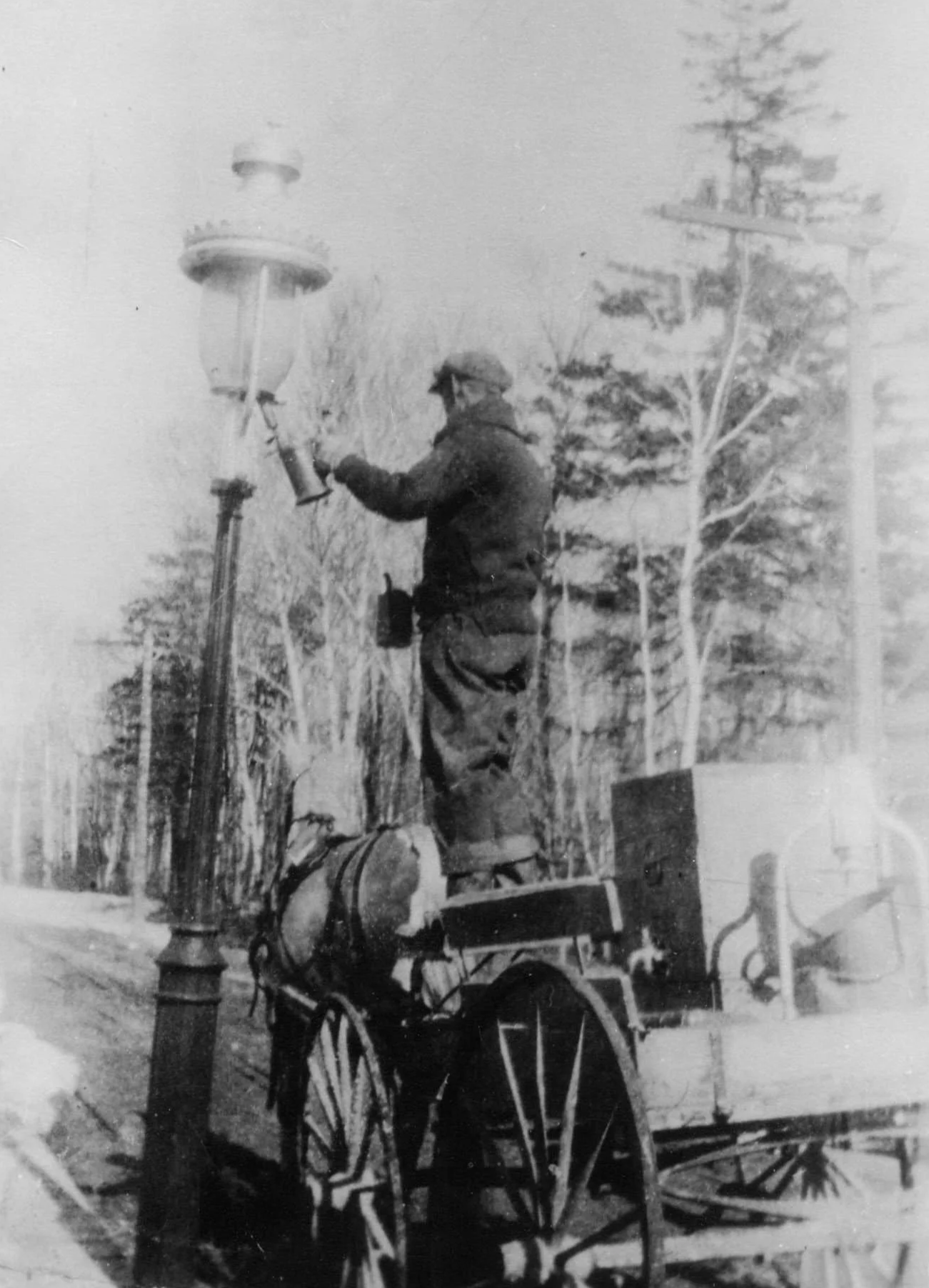 A black and white photo of a man in a coat, holding and working on a street lamp post next to a small horse-drawn carriage with equipment and a bag. There are trees and power lines in the background.