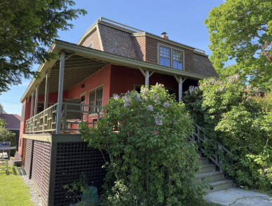 A two-story house with a red exterior, a front porch, and stairs leading up to it, surrounded by greenery and trees under a blue sky.