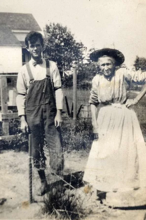 A young man and a woman standing outdoors in a garden, with a house and trees in the background. The man is wearing suspenders and a bow tie, and the woman is dressed in a long skirt and blouse, wearing a wide-brimmed hat.