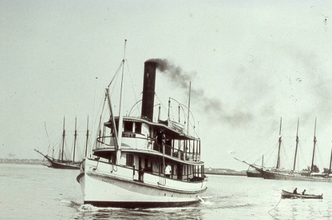 An old black and white photograph of a steamship with smoke coming out of its smokestack, sailing in the water with other ships in the background.