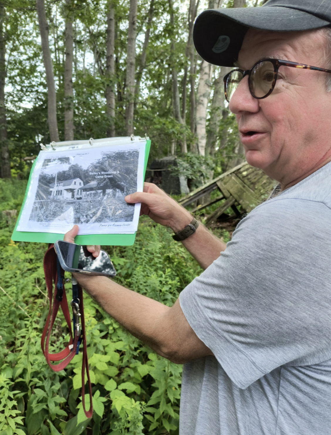 A man holding a photo album and a smartphone amidst green foliage in a wooded area, with trees and a dilapidated structure in the background.