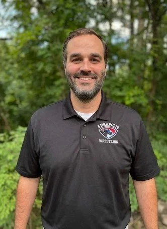 A smiling man with a beard and short hair, wearing a black polo shirt with an 'Annapolis Wrestling' logo, standing outdoors with green trees in the background.
