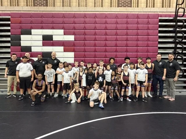 Group of young wrestlers and coaches posing in a gymnasium with pink and black bleachers in the background.