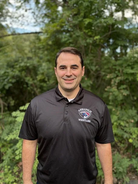 Man standing outdoors in front of green trees, wearing a black Annapolis Wrestling polo shirt, smiling at the camera.