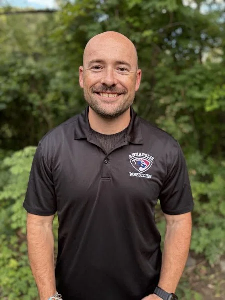Man with a beard smiling, wearing a black polo shirt with a logo, outdoors with trees in the background.