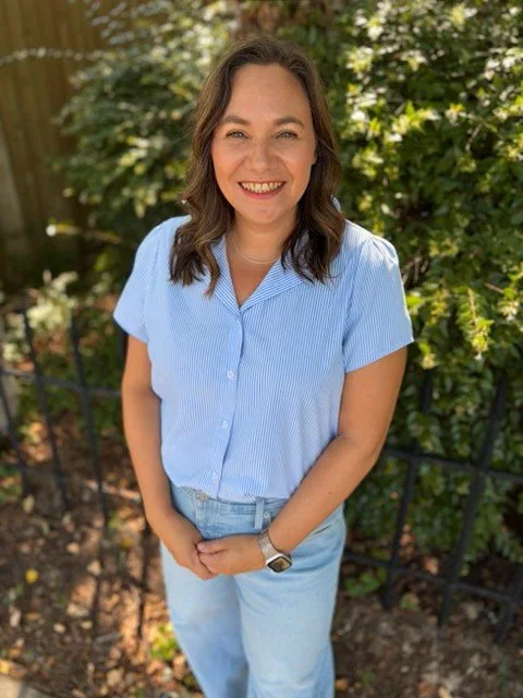 A woman with shoulder-length dark hair smiling outdoors, wearing a light blue short-sleeved button-up shirt and jeans, standing in front of green foliage.