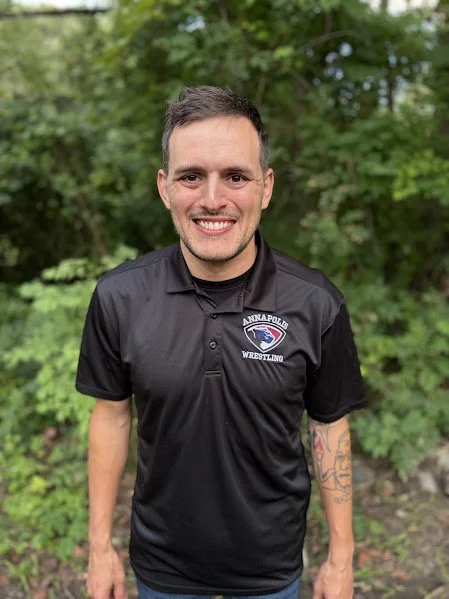 A man standing outdoors in front of green foliage, wearing a black Annapolis Wrestling polo shirt, smiling at the camera.