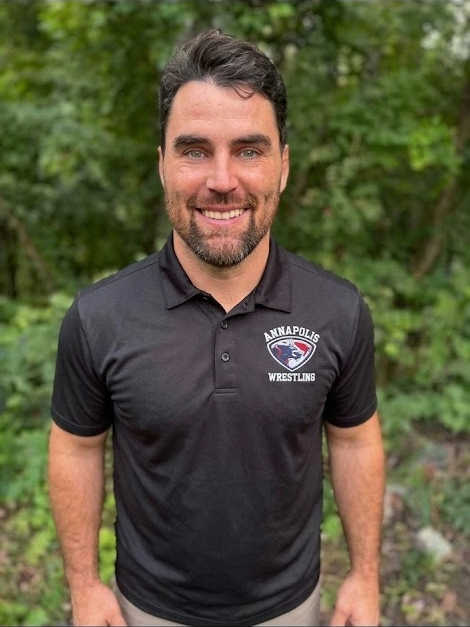 Man smiling outdoors wearing a black polo shirt with 'Annapolis Wrestling' logo, green trees in background.