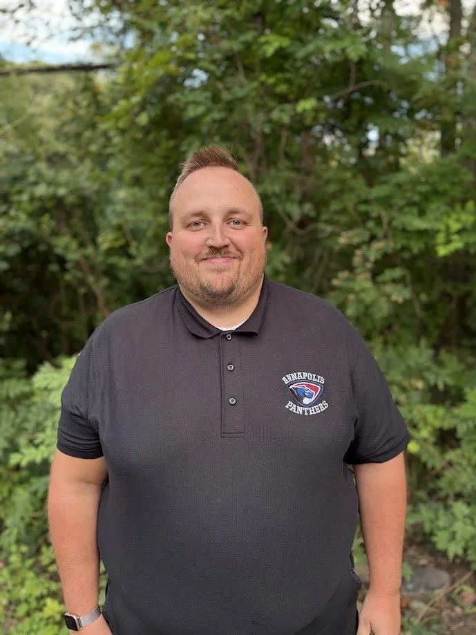 A man with short hair and a beard, smiling and standing outdoors in front of green foliage, wearing a black polo shirt with an 'Annapolis Panthers' logo on the left chest.