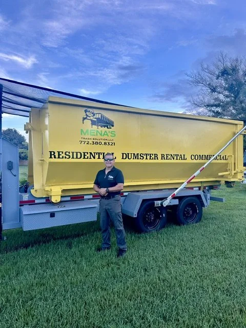 A man standing in front of a large yellow dumpster with the logo for MENA'S Trash Solutions, located outdoors on a grassy area under a partly cloudy sky.