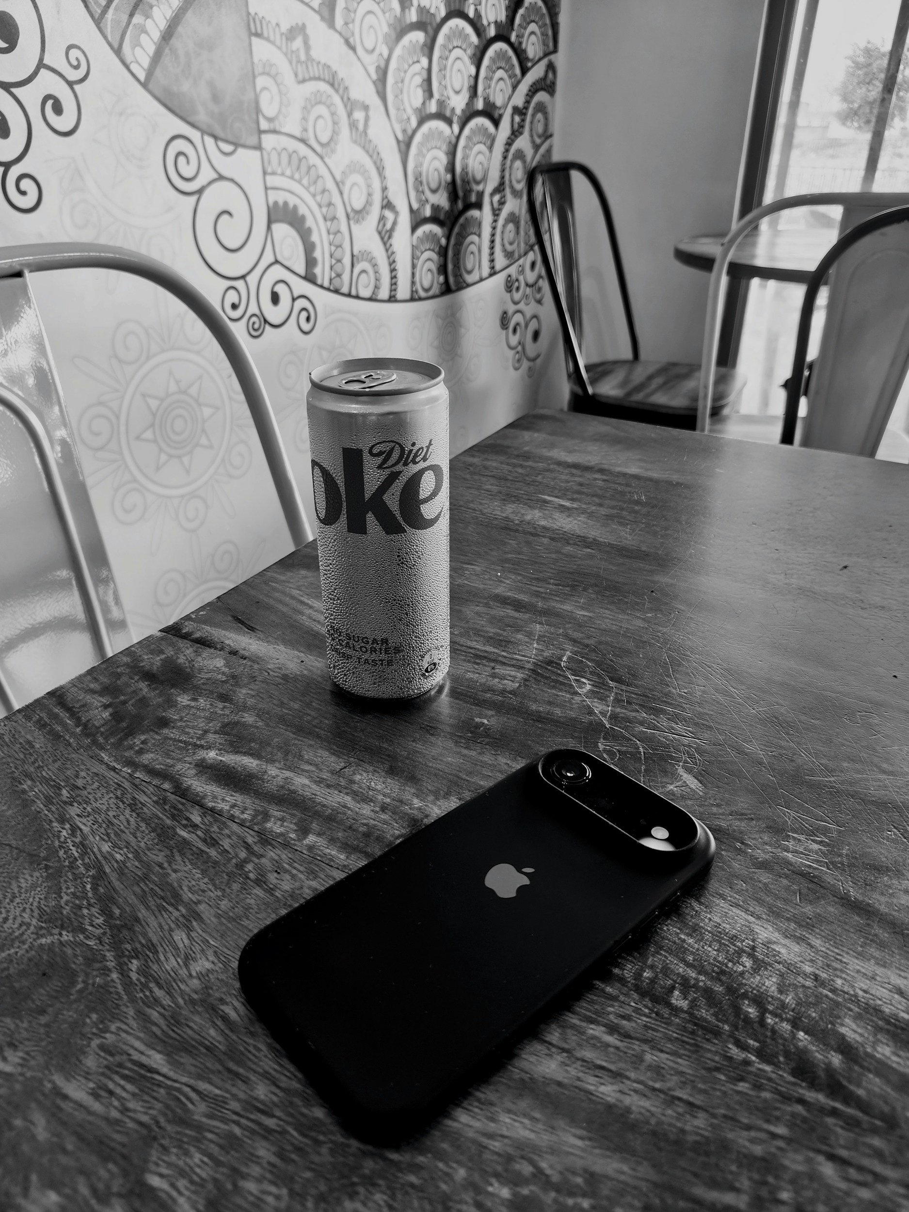 A desk with a Diet Coke can, a smartphone, and a laptop in a cafe with patterned wall art and chairs near a window.
