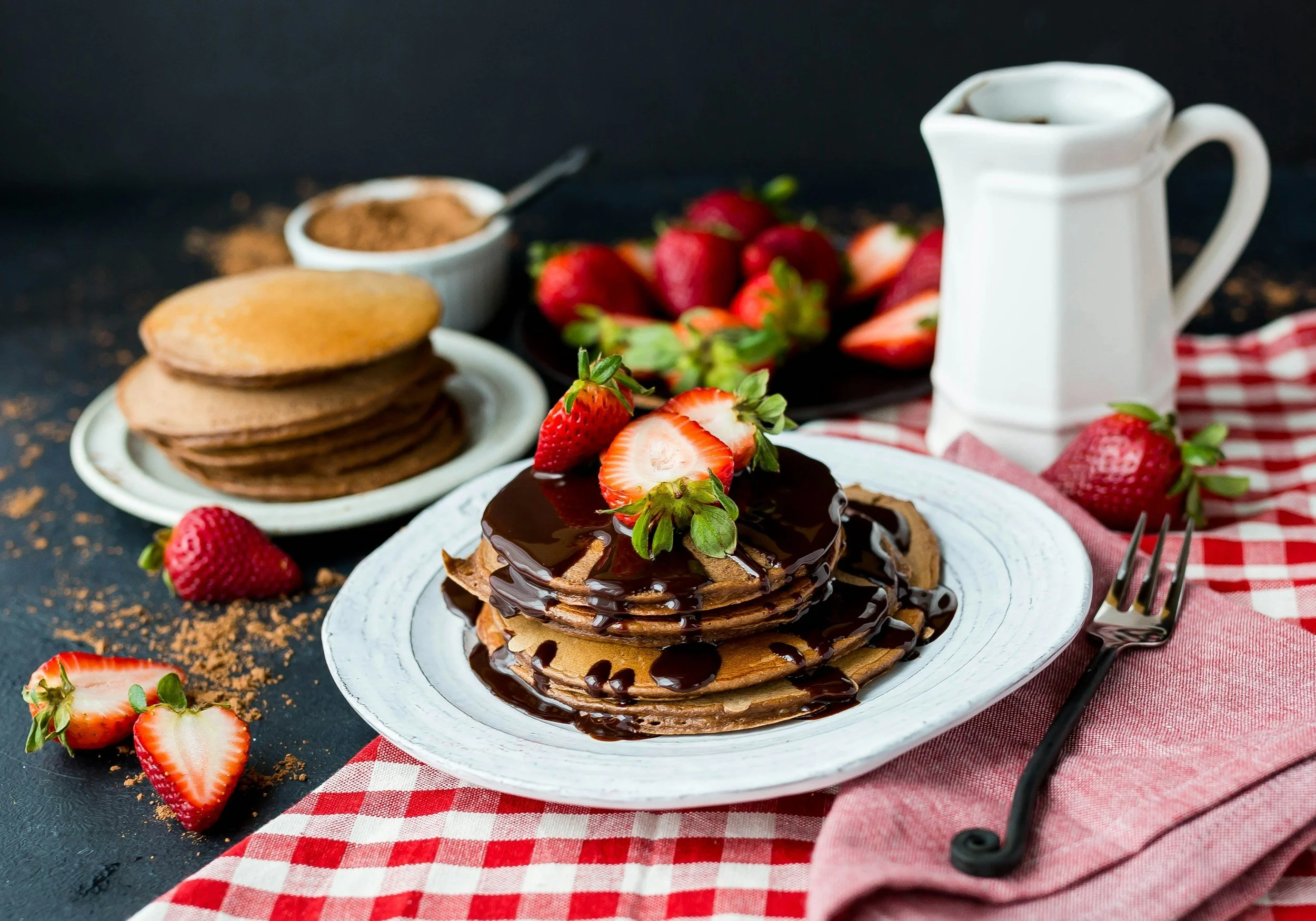 Stacked chocolate pancakes topped with strawberries and chocolate syrup on a white plate, with additional strawberries and a jar of syrup in the background, on a dark surface with a red and white checkered cloth.