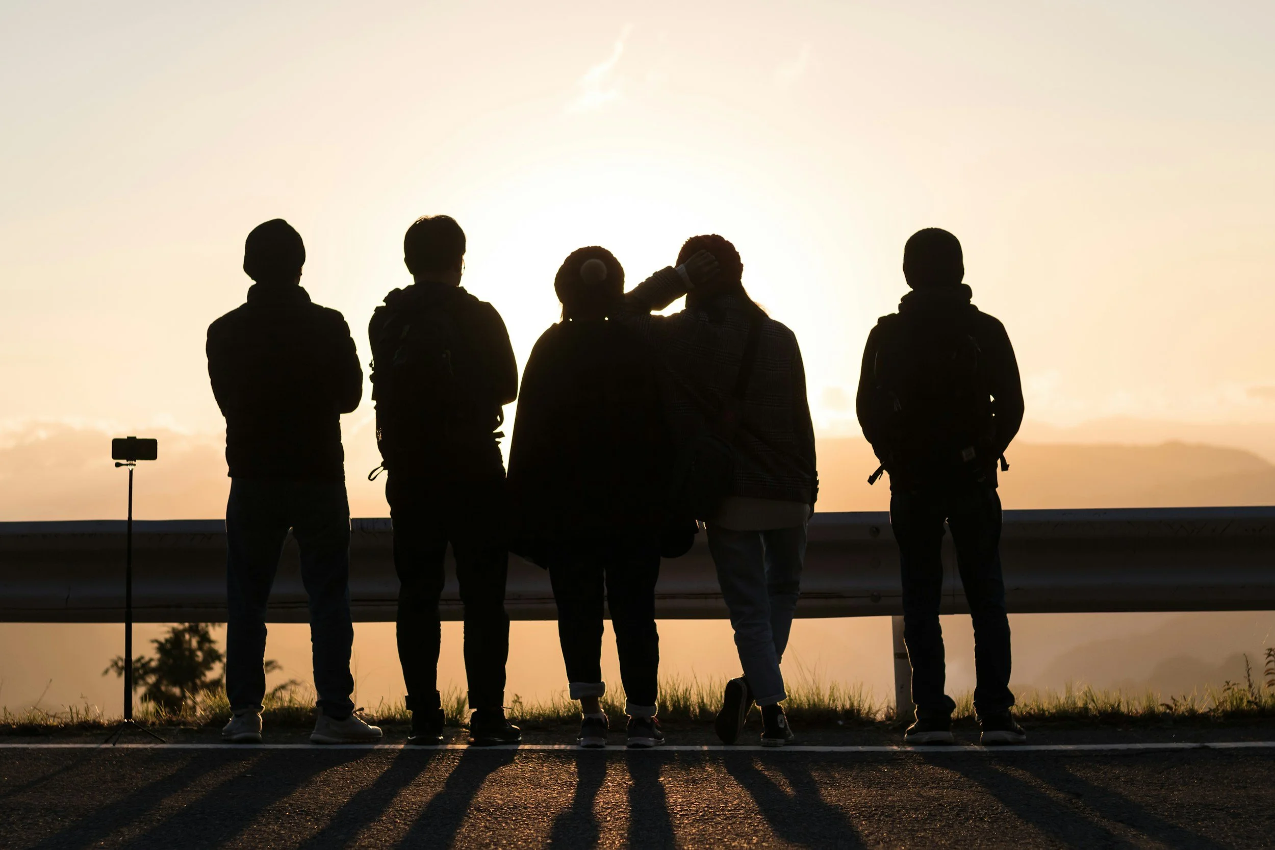 Five people standing near a guardrail at sunset, with one person photographing the scene. The group appears to be enjoying the view as the sun sets behind them.