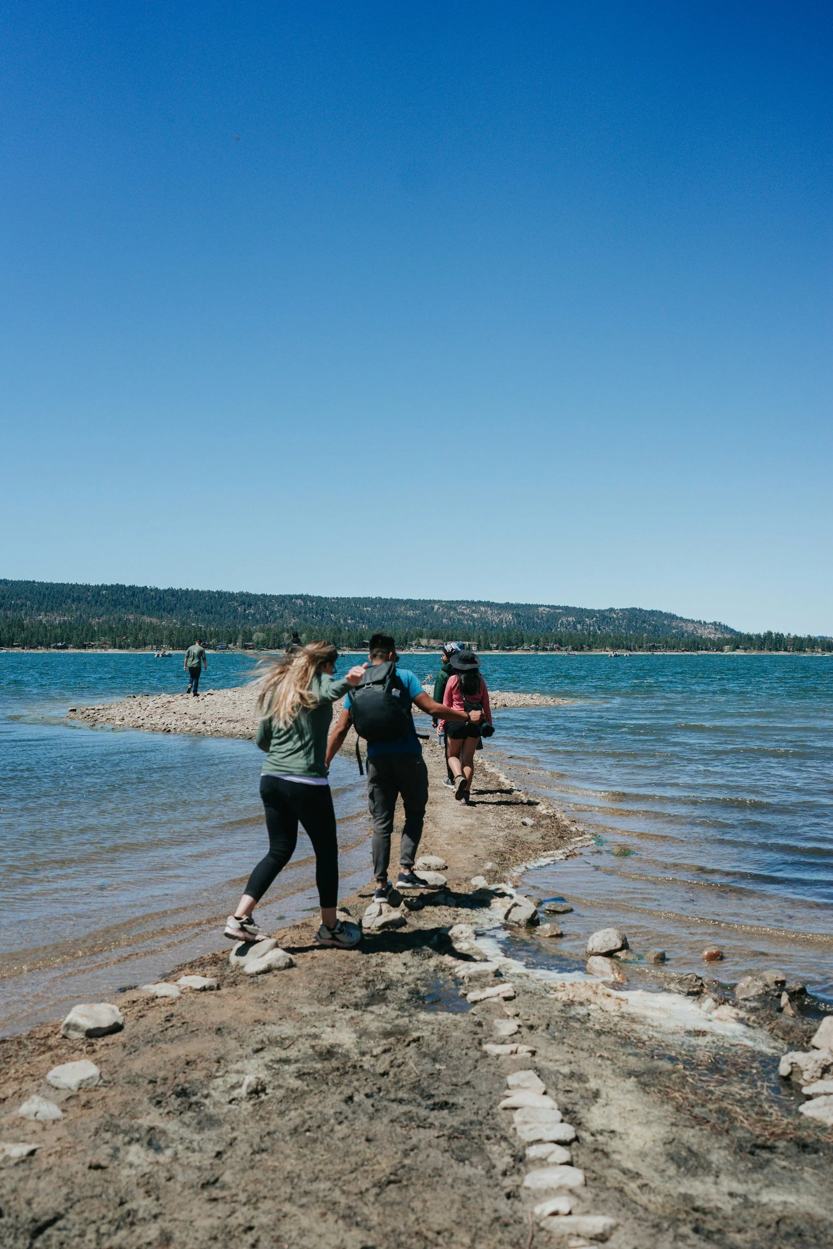 People hiking or walking along a small natural stone path into a lake on a sunny day with a forested hill in the background.