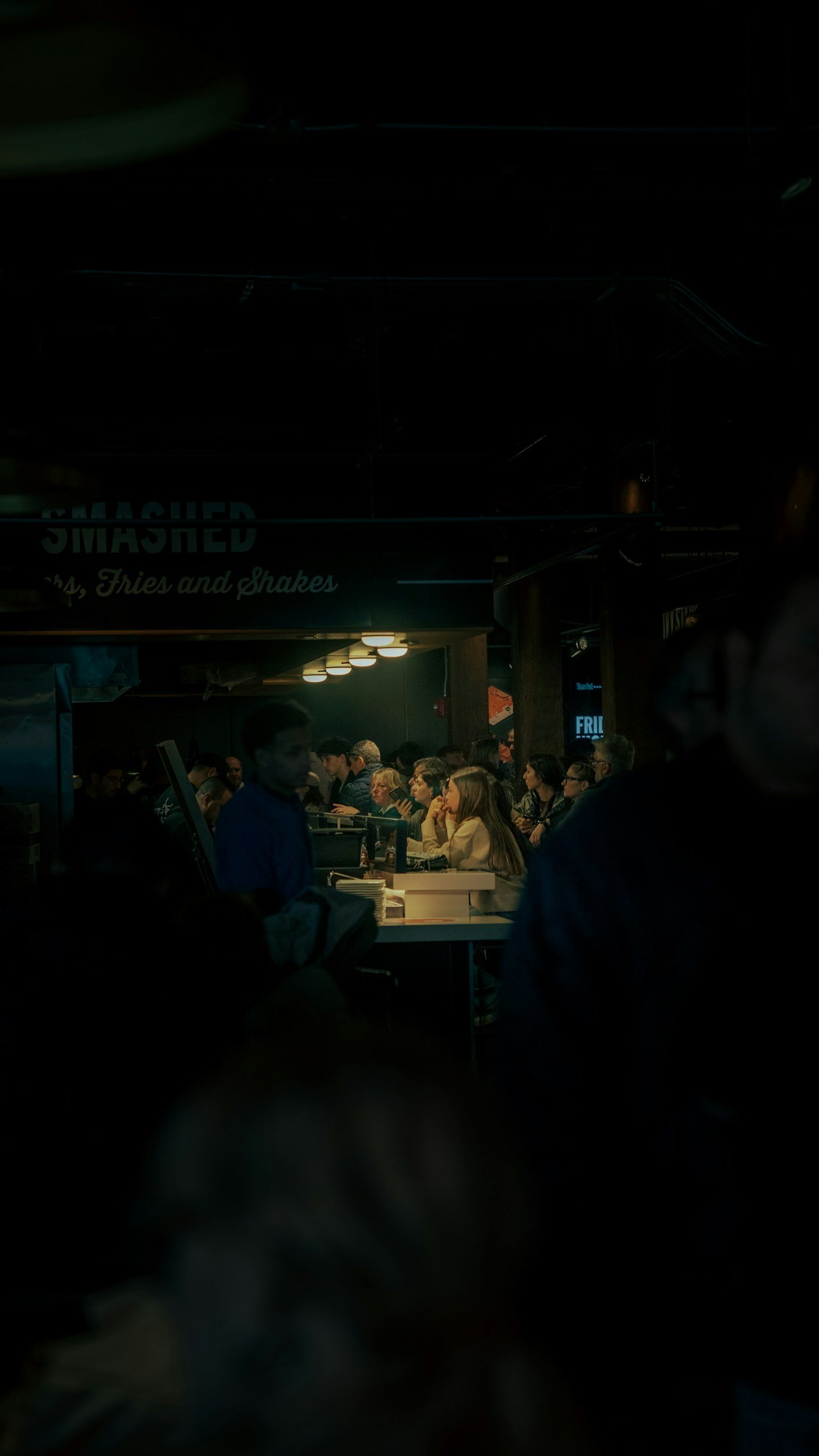 A busy indoor scene at a food counter with several people waiting and ordering food. The background has a sign that reads 'Smashed' and mentions fries and shakes. There are multiple seated diners and a staff member at the counter.