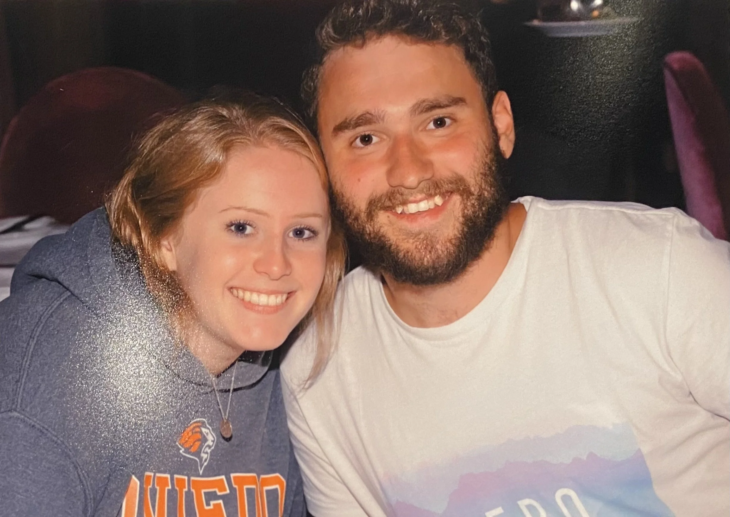 A young woman with red hair and a young man with dark, curly hair and a beard, smiling and posing closely together in a dimly lit setting with a dark background.