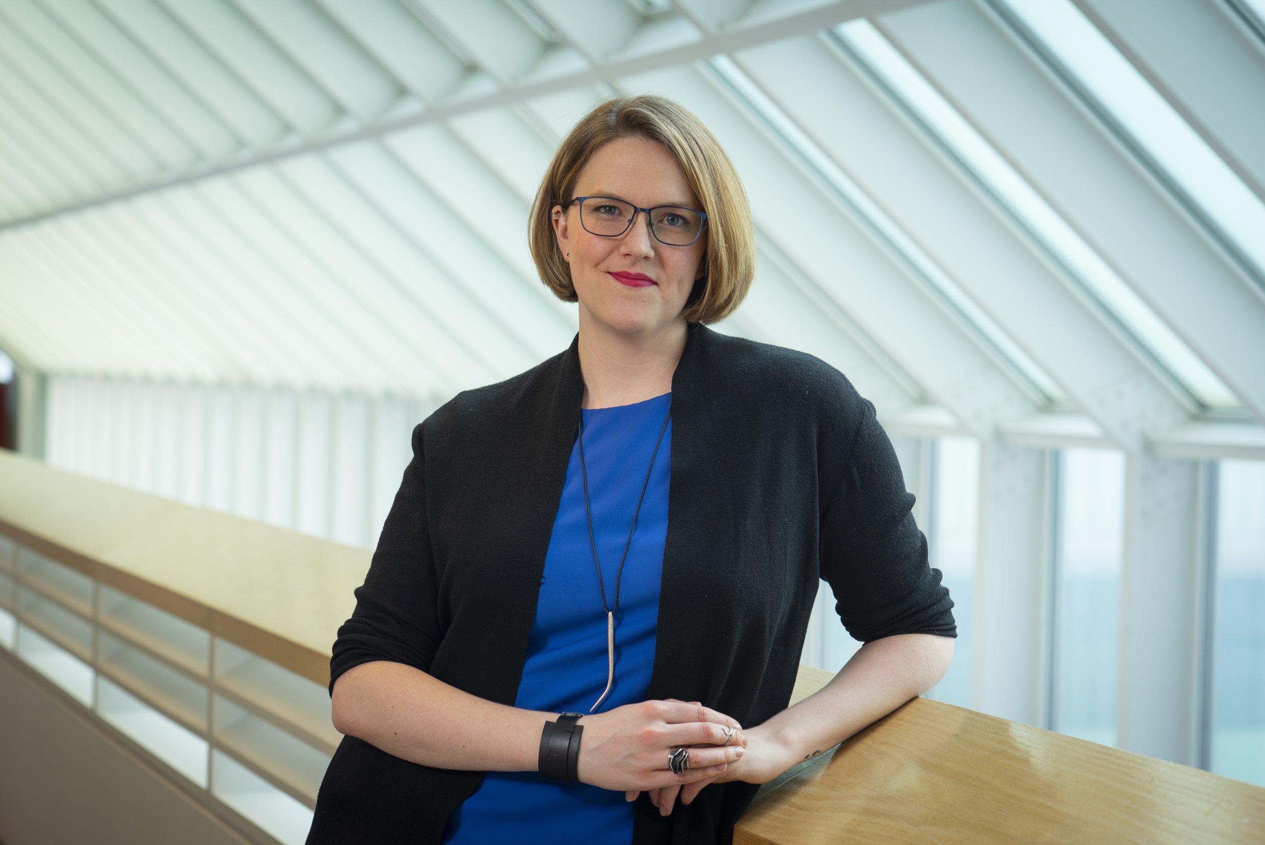 A woman with short blonde hair, glasses, and red lipstick, wearing a black blazer over a blue top, leaning on a wooden railing in a modern building with large windows and a glass ceiling.