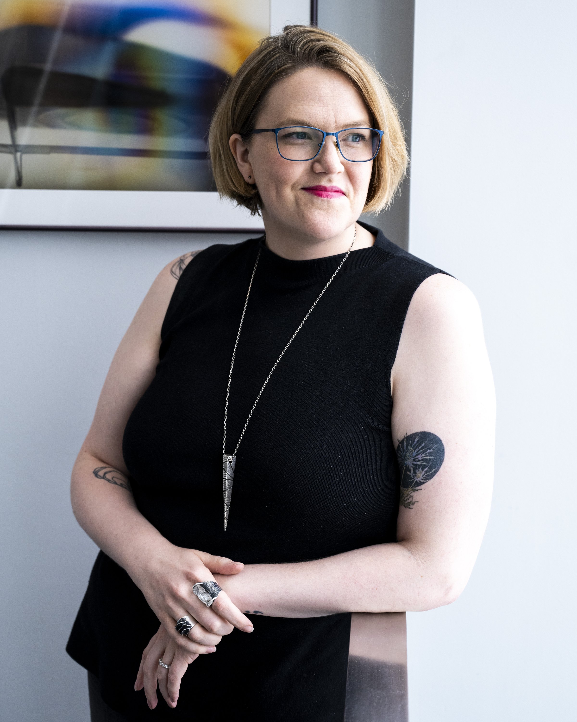 A woman with short, light brown hair, wearing glasses with blue frames, a black sleeveless top, and jewelry, stands with her arms crossed and looks to her left, standing in front of a colorful abstract painting and a white wall.
