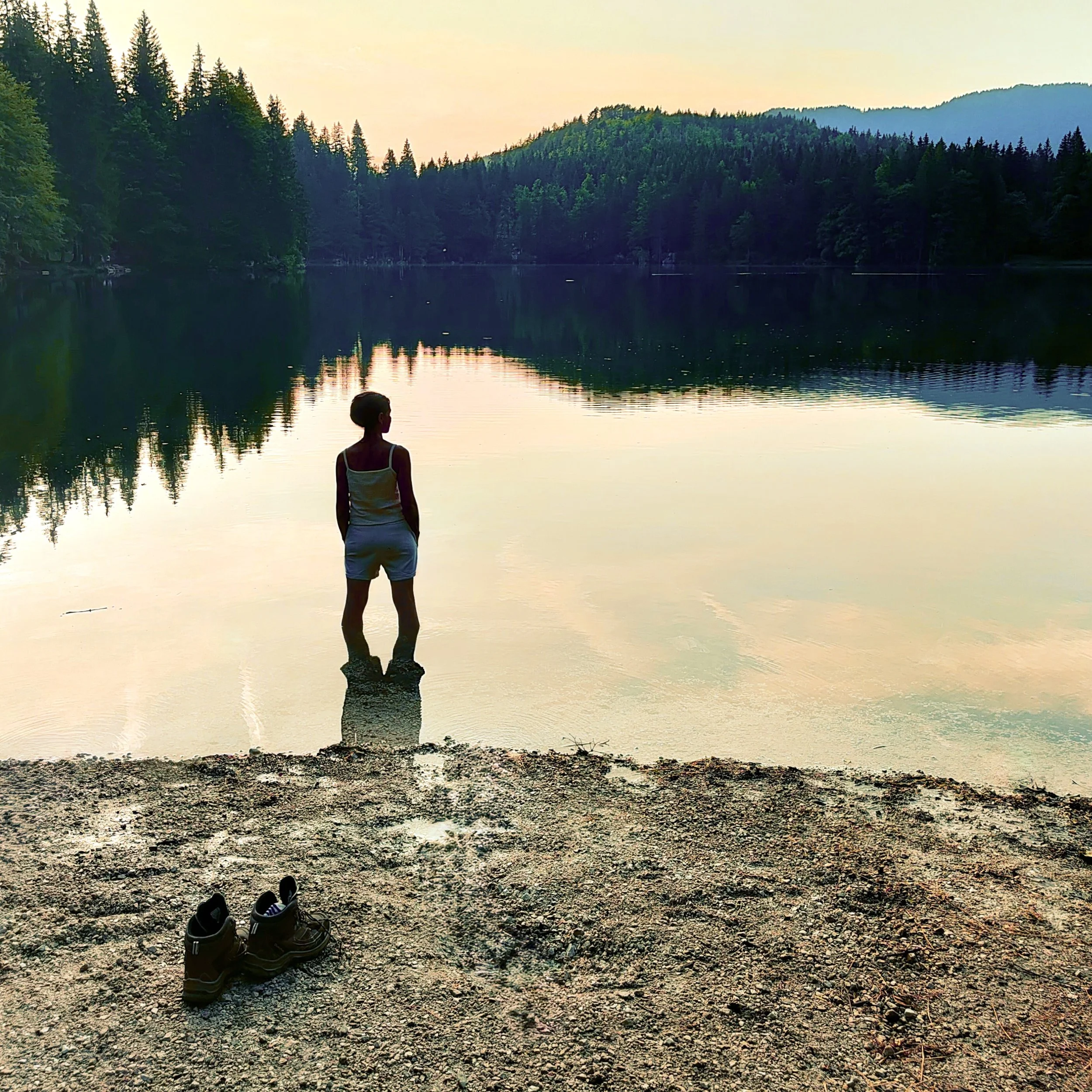 Ragazza che si trova davanti a un lago circondato da alberi e montagne, con scarpe poste sul terreno roccioso in primo piano.