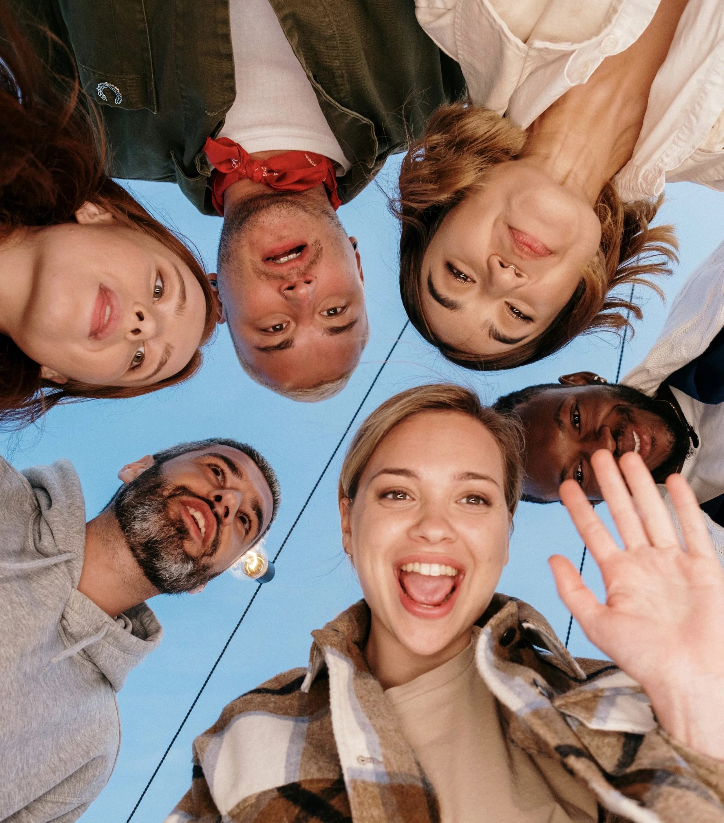 Un gruppo di persone sorridenti si riuniscono in cerchio e si scattano una foto dall'alto, con un cielo azzurro sullo sfondo.