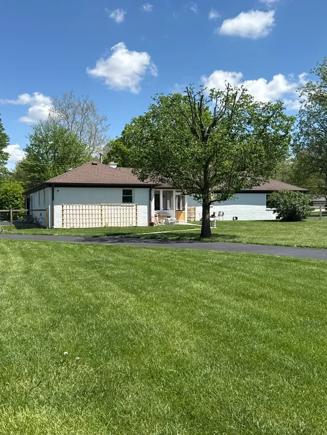 A white house with a brown roof and a fenced backyard, surrounded by green grass and trees under a blue sky with white clouds.