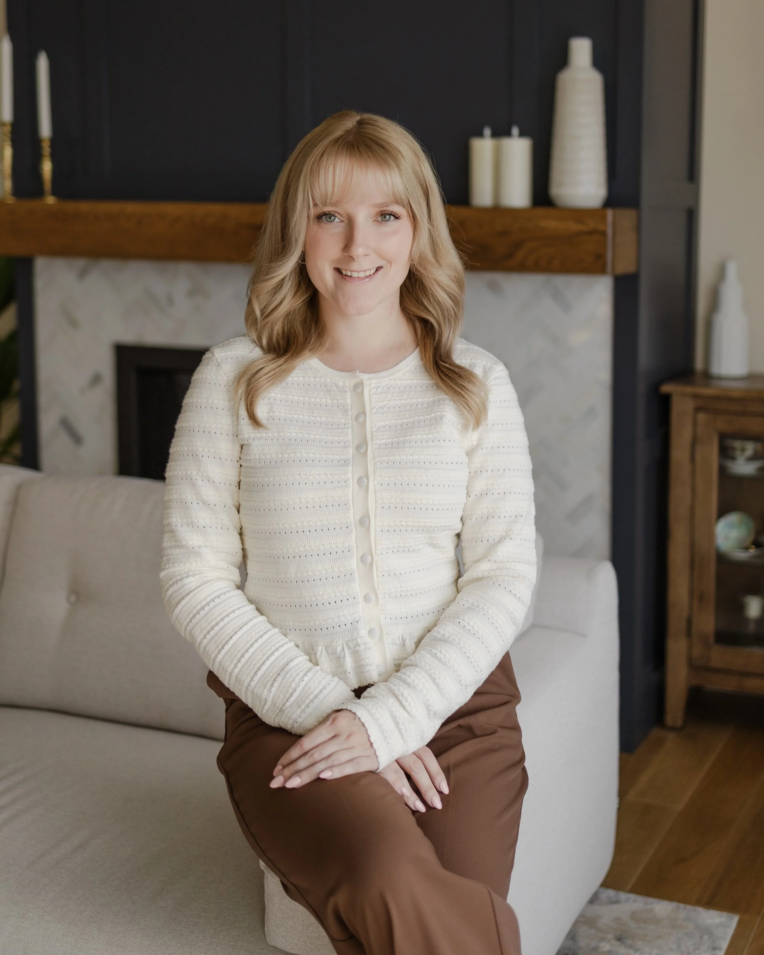A woman with blonde hair, wearing a cream-colored cardigan and brown pants, sitting on a beige sofa in a living room with a fireplace and decorative vases.