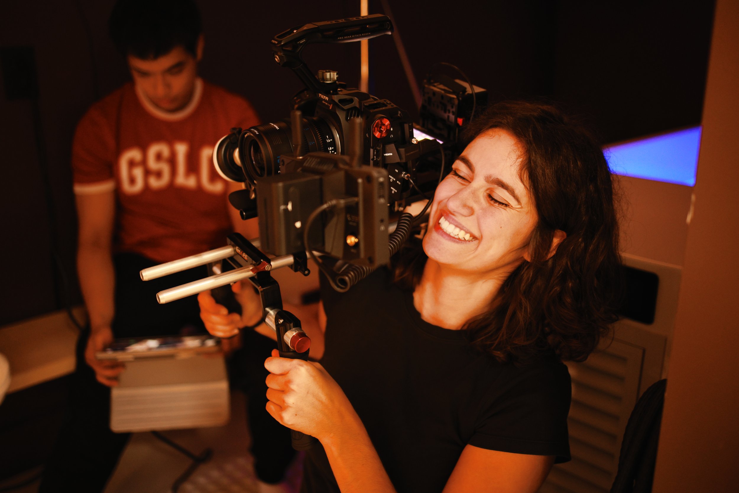 Woman smiling while holding a camera rig during filming. Man in background adjusting equipment in a dimly lit room.