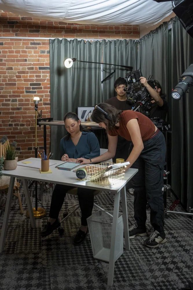A woman is sitting at a desk with notebooks and a plant, while a researcher leans over to make notes or adjustments, surrounded by filming equipment and two camera operators in a studio with brick walls and green curtains.