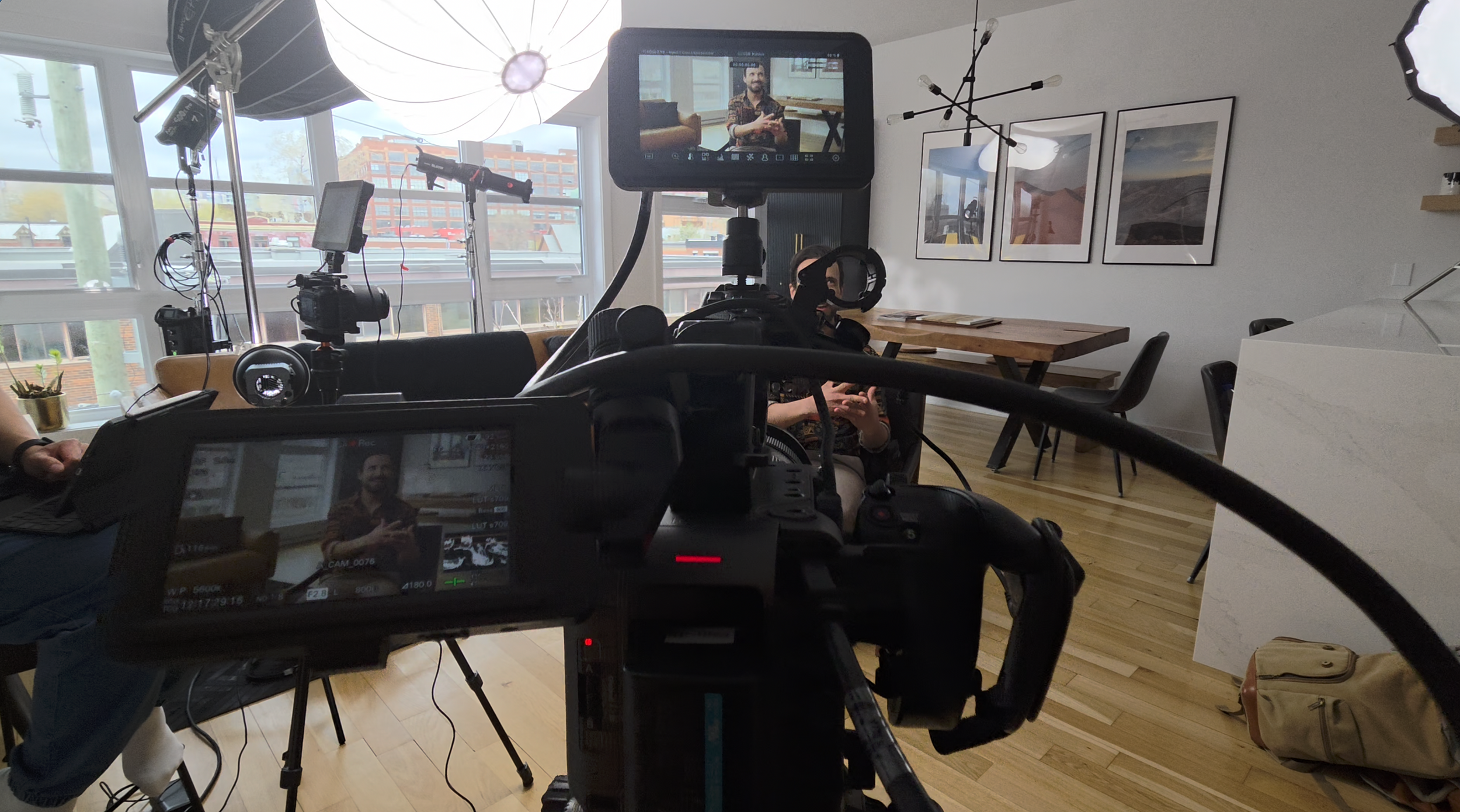 A camera recording a person during an indoor interview or video shoot with filming equipment, lights, and framed pictures on the wall.