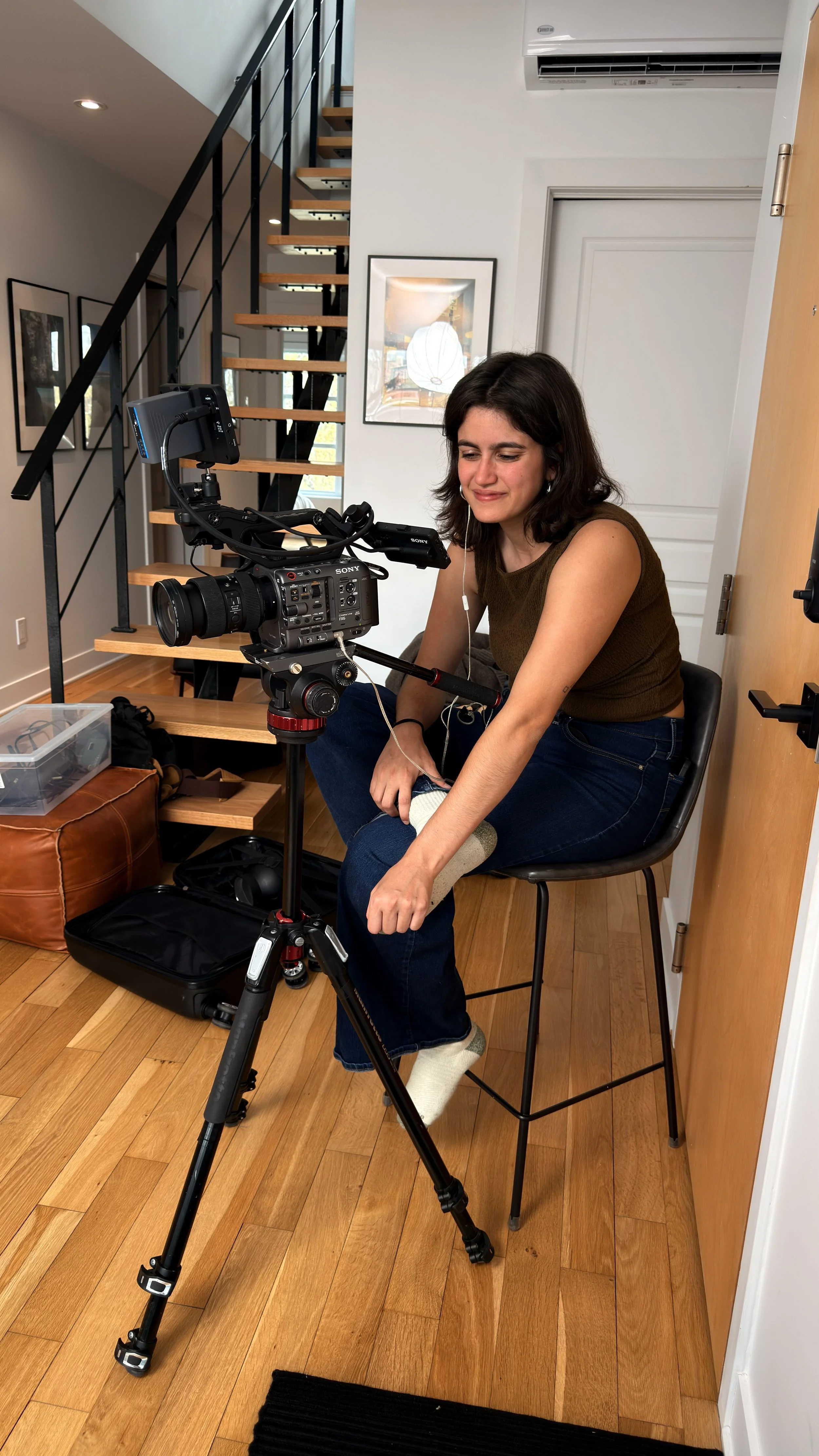 A woman sitting on a chair placing a sock on her foot while filming with a professional camera on a tripod inside a home with wooden flooring, staircase, and framed pictures on the wall.
