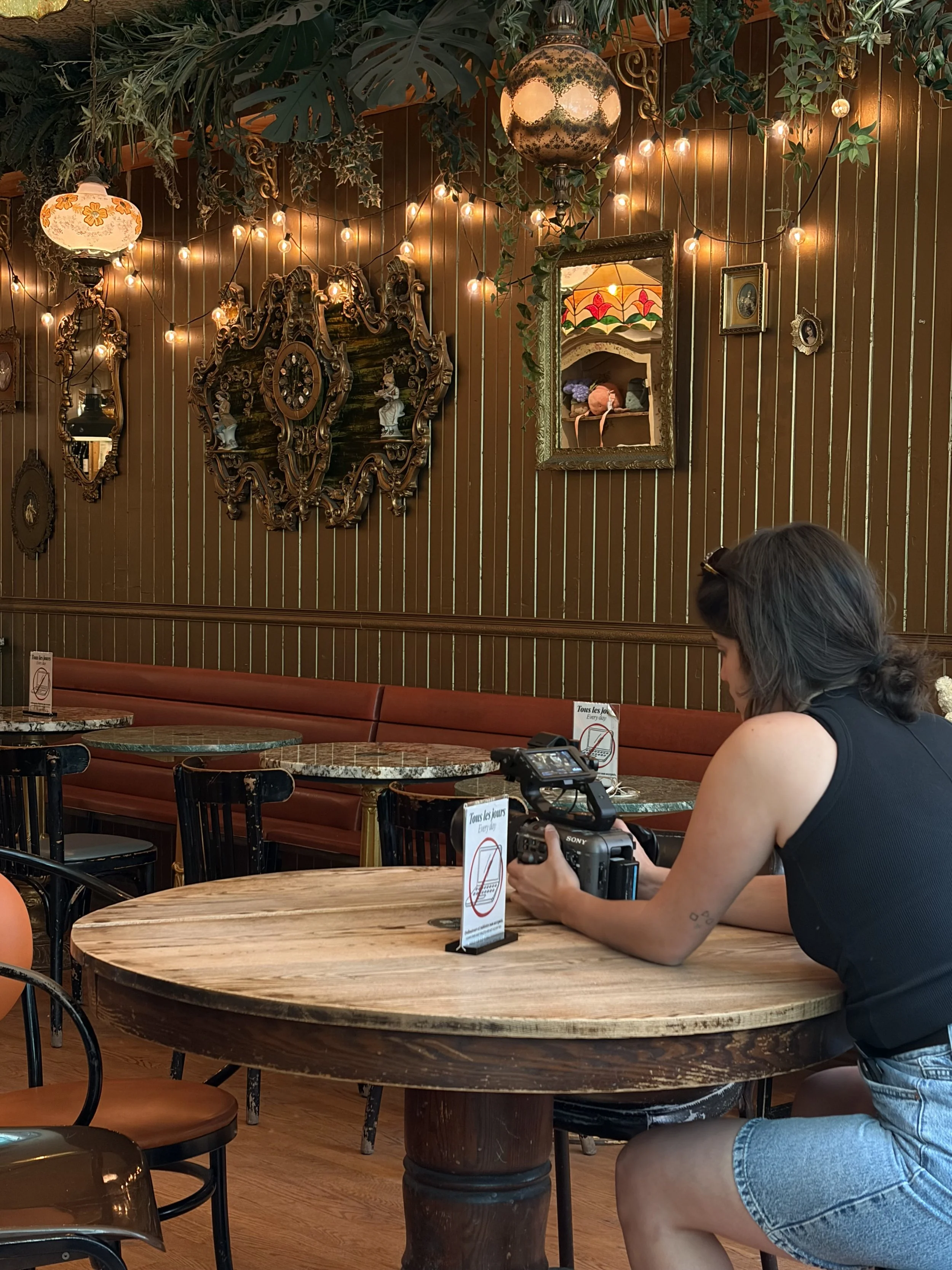 A woman in a black tank top and denim shorts sitting at a round wooden table in a cozy cafe, holding a camera, with vintage-inspired lighting and ornate wall decorations behind her.