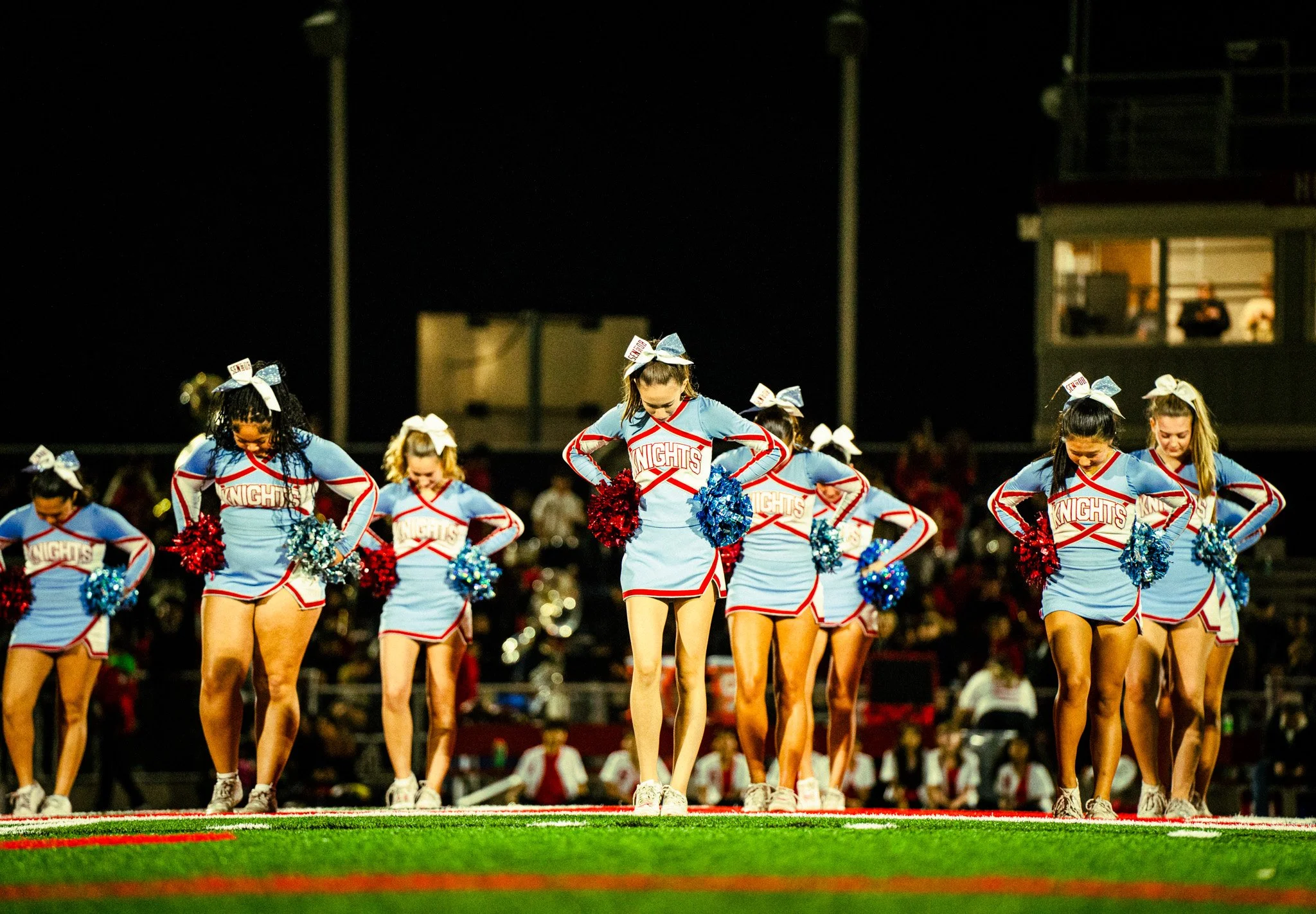 Cheerleaders performing on a football field at night, wearing blue and red uniforms with large bows in their hair.