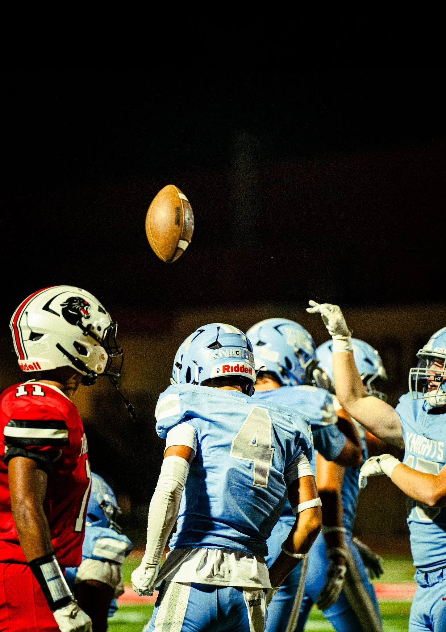 Football game at night with players in blue and red uniforms and helmets, one player throwing a football.