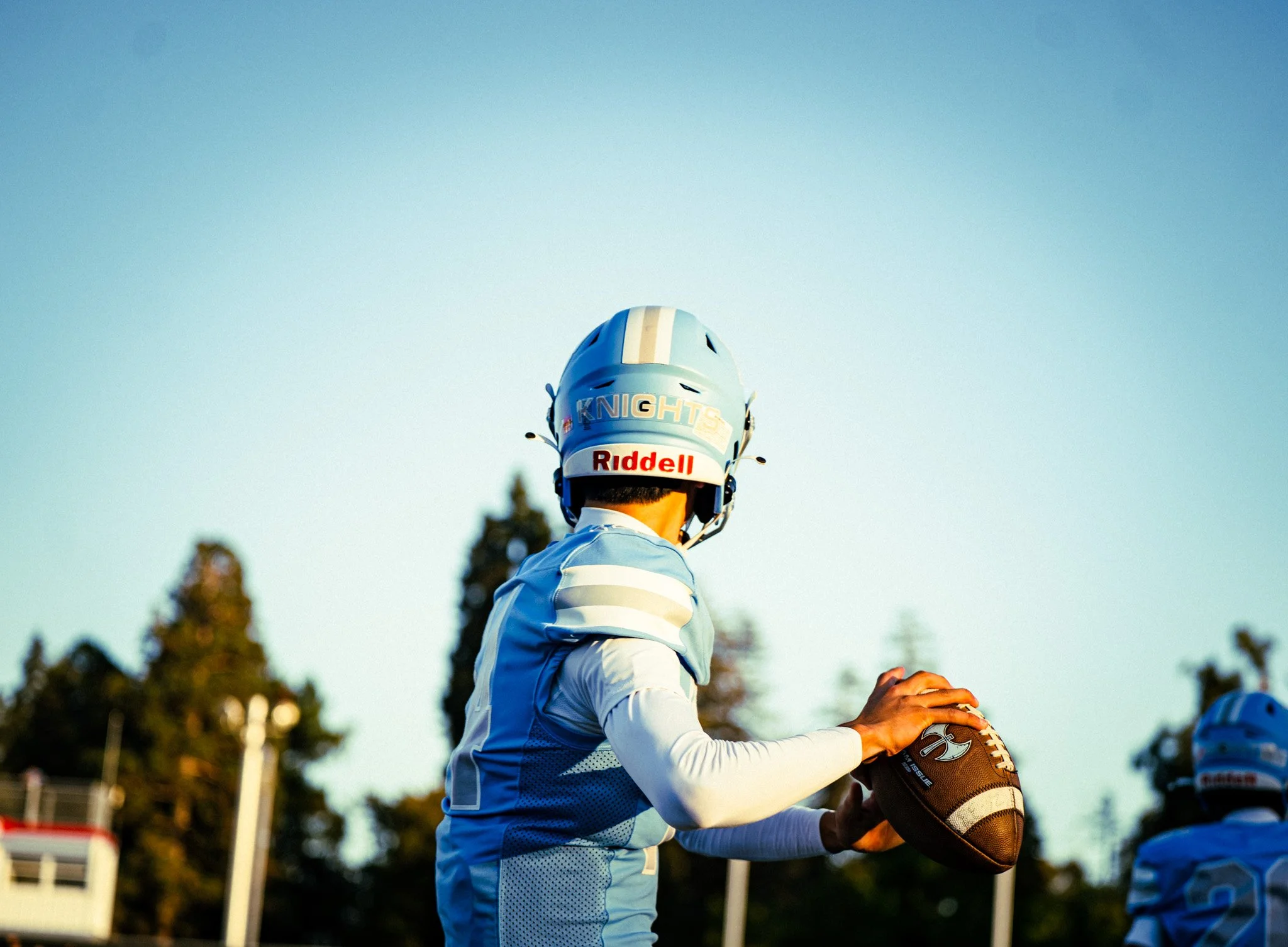 A football player wearing a gray and blue uniform and a helmet labeled "KNIGHTS" is holding an American football, preparing for a play on a football field during daytime.