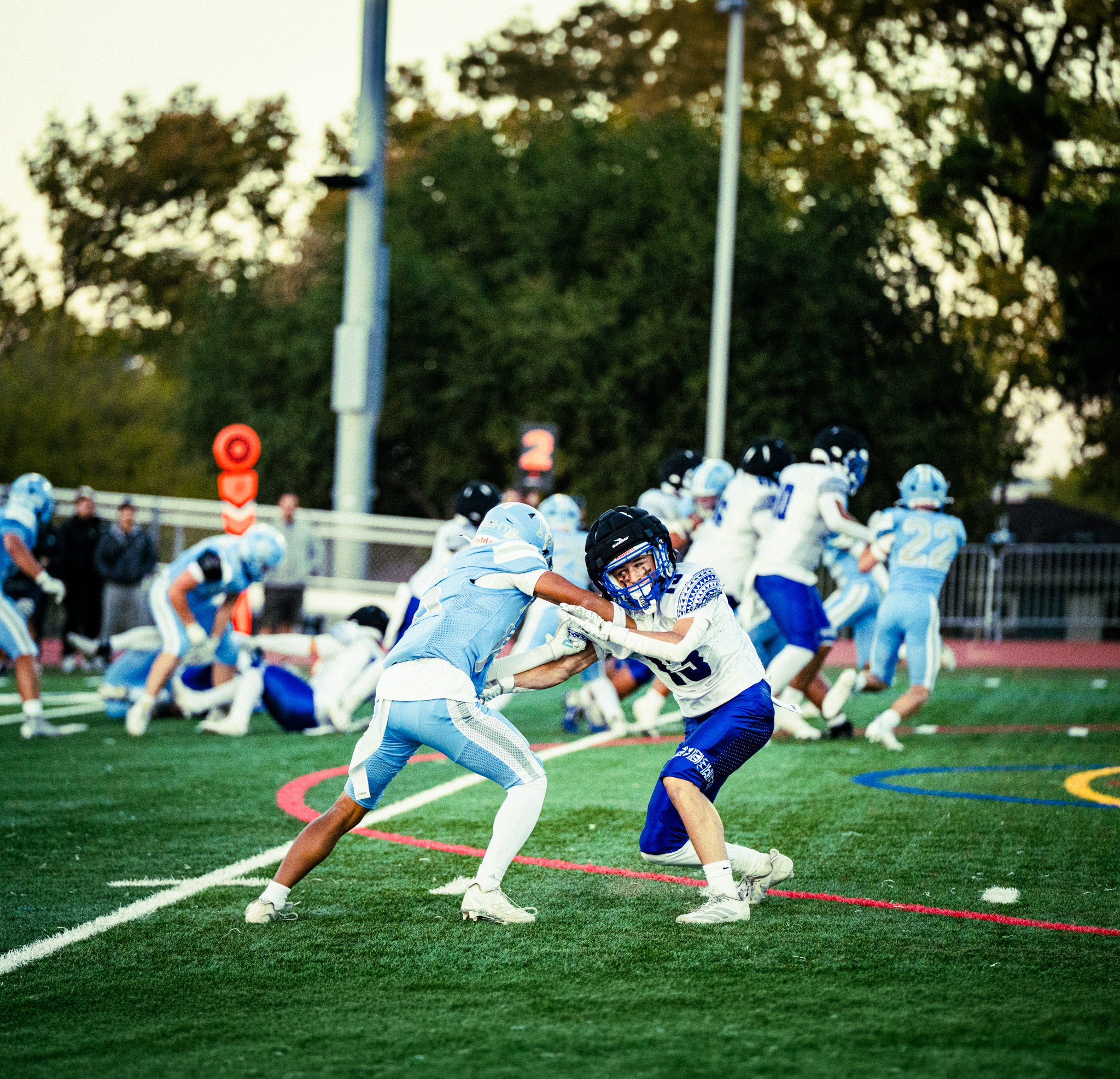 Two young football players gripping each other's jerseys during a game on a green field, with other players and spectators in the background. Trees and a metal fence are visible beyond the playing area.